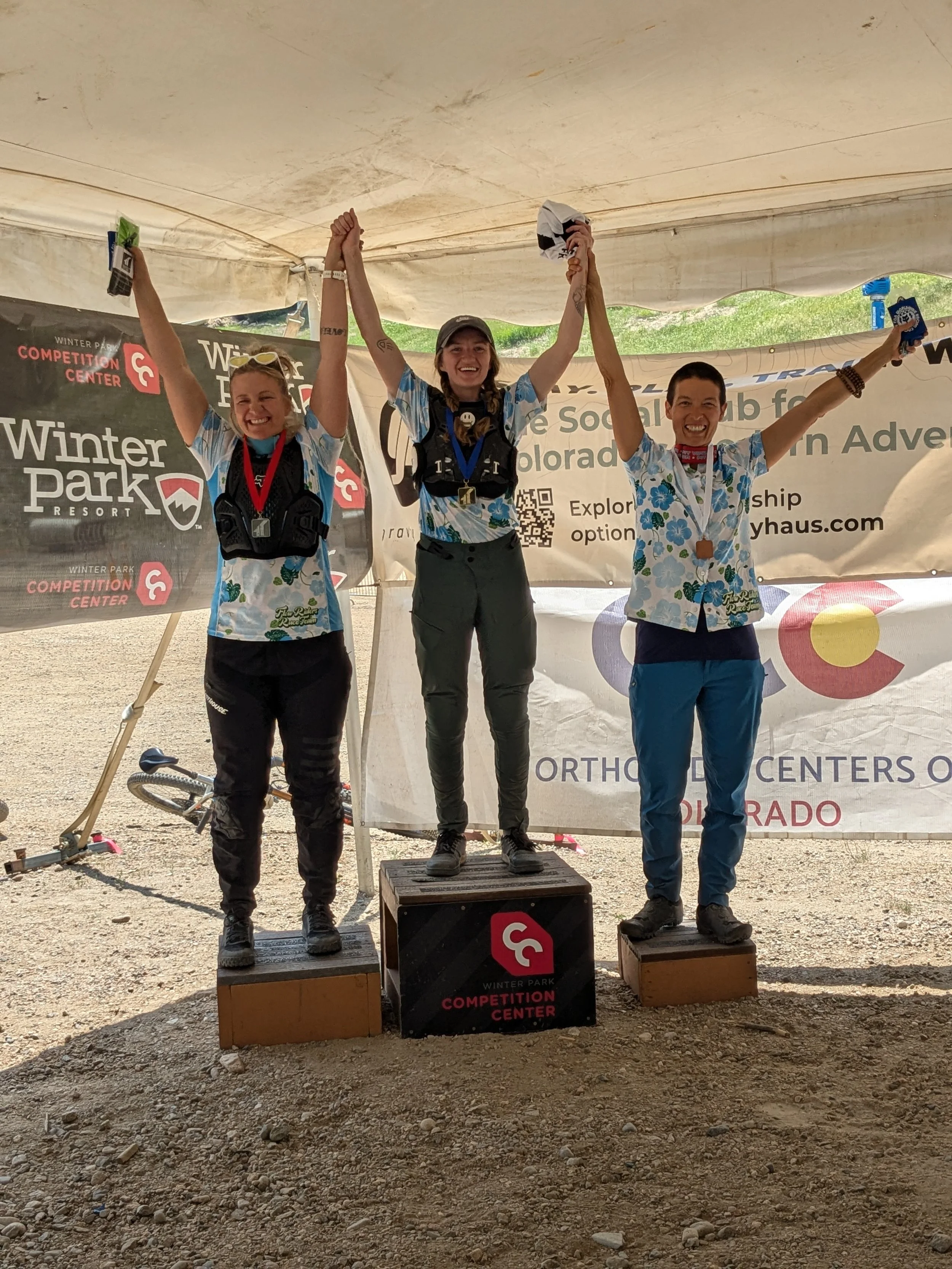 Three women standing on winners' podiums at a competition, holding their arms up in victory. The woman in the center is on the highest podium, wearing a black cap and black pants. The woman on the left is on a slightly lower podium, wearing black pants and sunglasses on her head. The woman on the right is on the third-place podium, wearing blue pants. They are all wearing medals and have smiles on their faces.