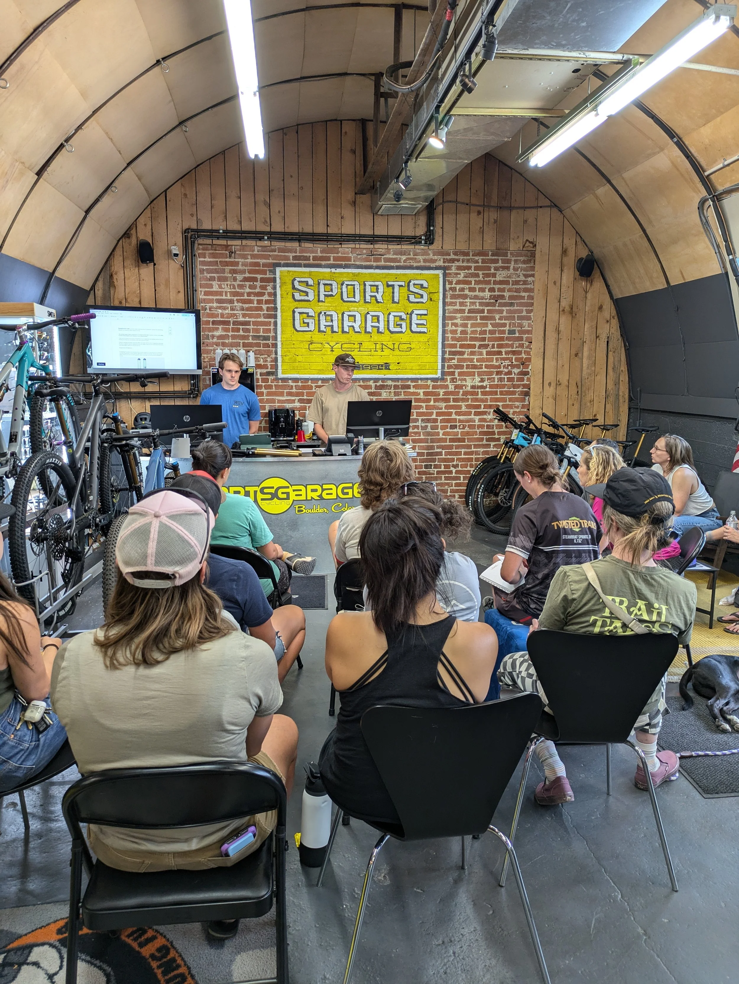 People attending a presentation at Sports Garage cycling store with a brick wall background and bicycles displayed.