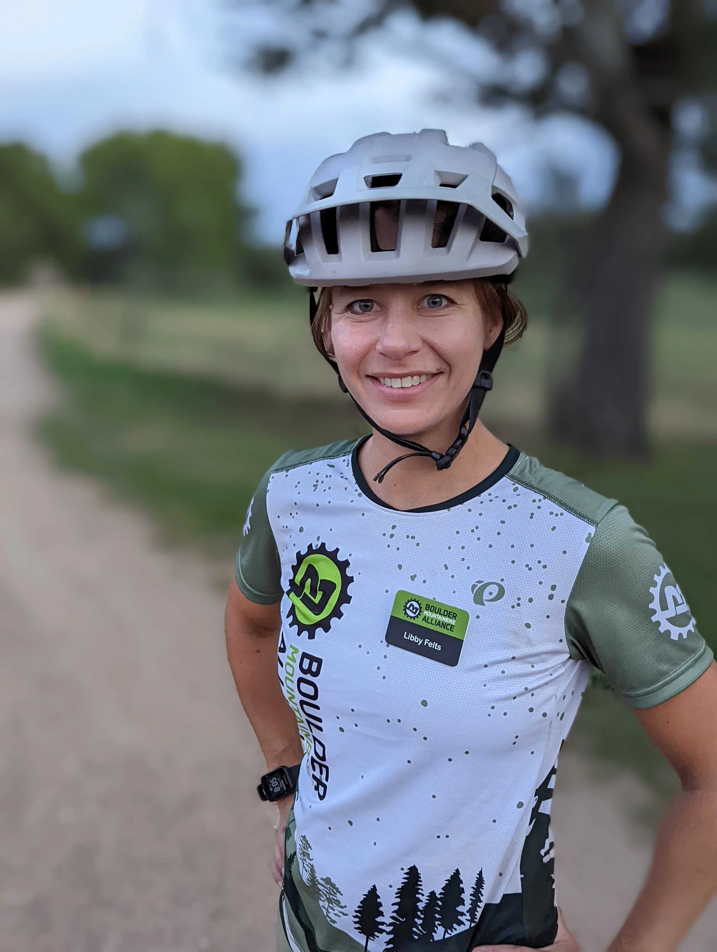 Woman with short hair wearing a bike helmet, sports shirt with outdoors and mountain bike graphics, smiling outdoors on a dirt path with trees in the background.