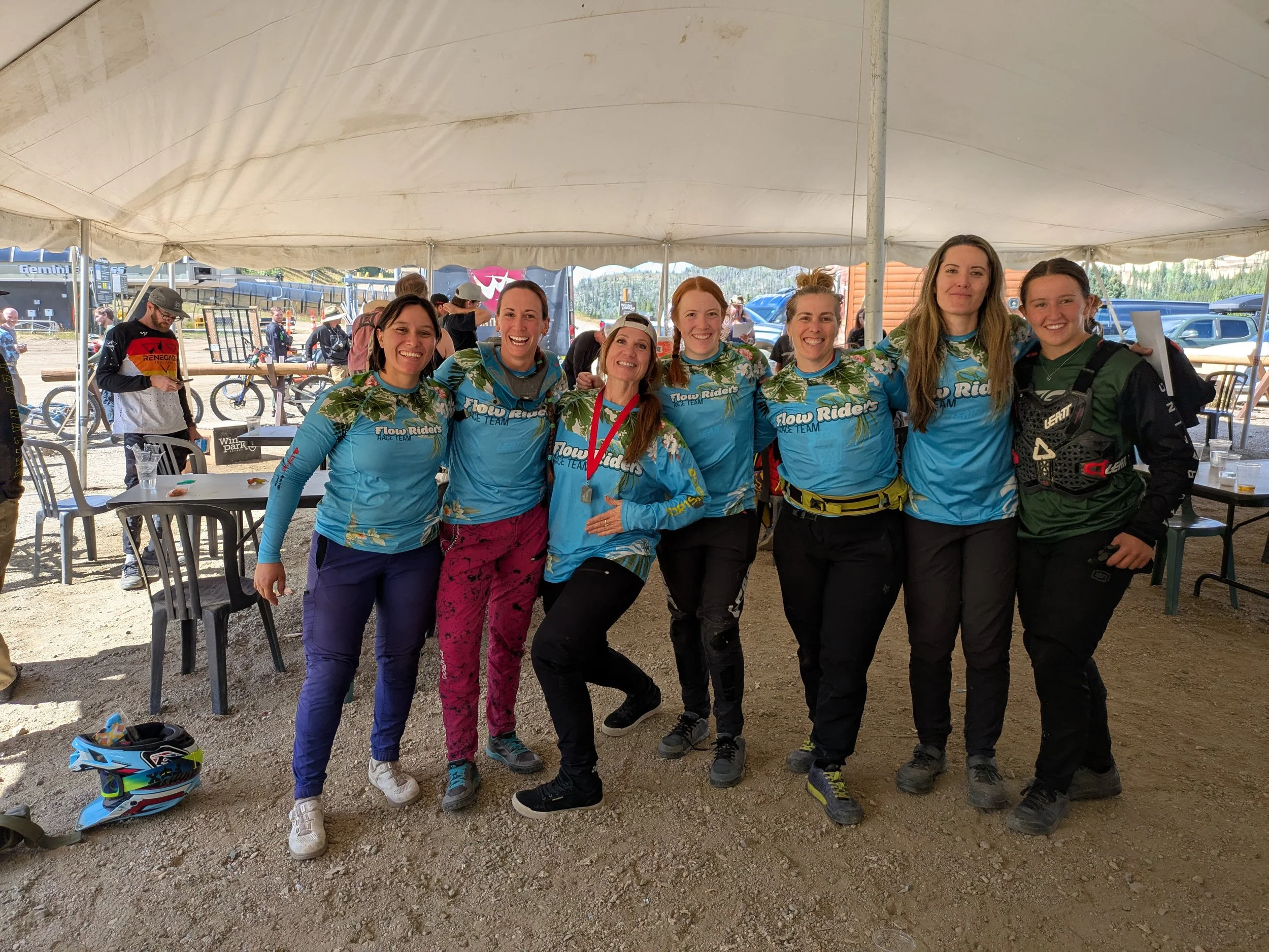Group of eight women standing together under a tent, smiling, with some wearing matching blue Fox Ride jerseys. One woman in the middle is with a medal around her neck. The background shows other people, bicycles, and parked cars.