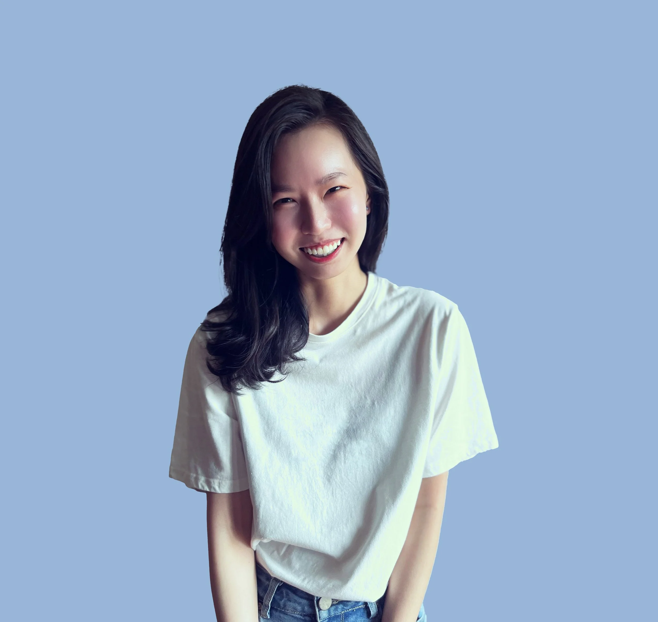 A young woman with long dark hair smiling and winking, wearing a white t-shirt and jeans, standing against a light blue background.