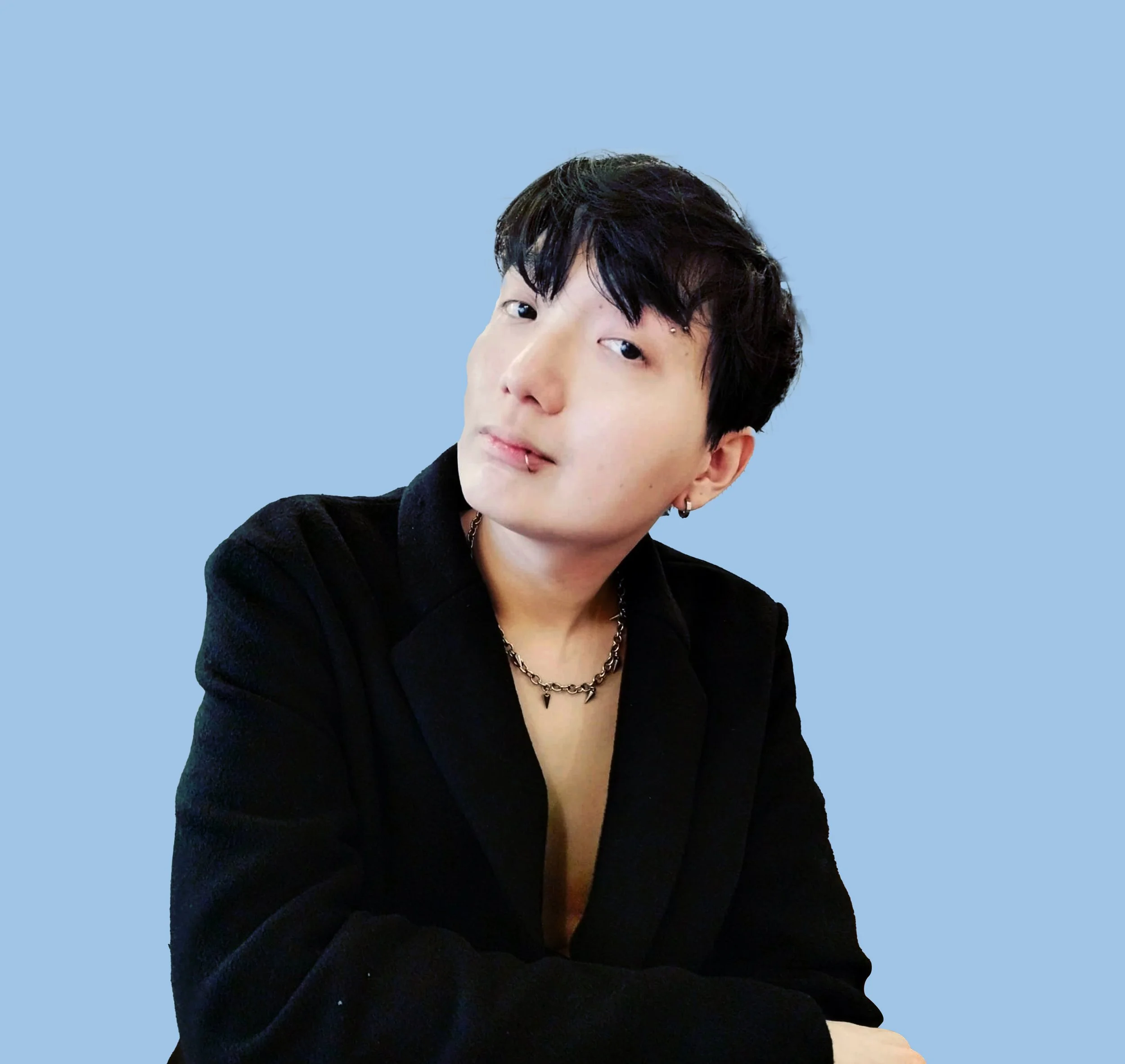 A young person with black hair, earrings, and a lip piercing, wearing a black jacket and chain necklace, posing against a blue background.