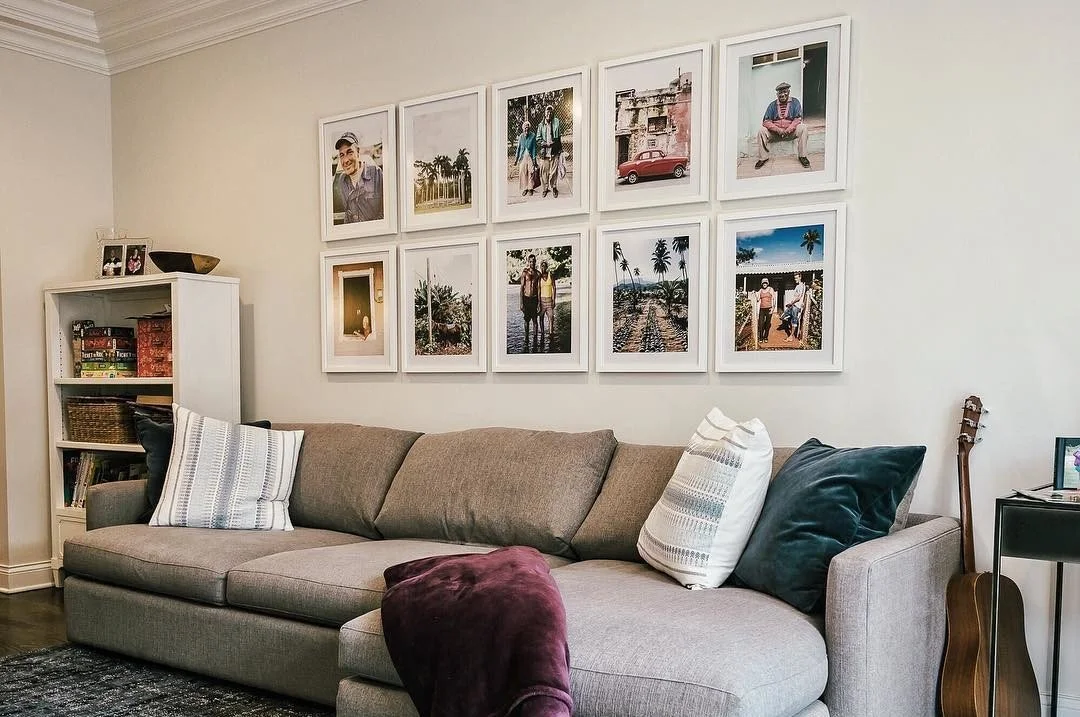 Living room with a beige couch adorned with three decorative pillows, artwork on the wall, a white bookshelf filled with books on the left, and a guitar propped against the wall on the right.