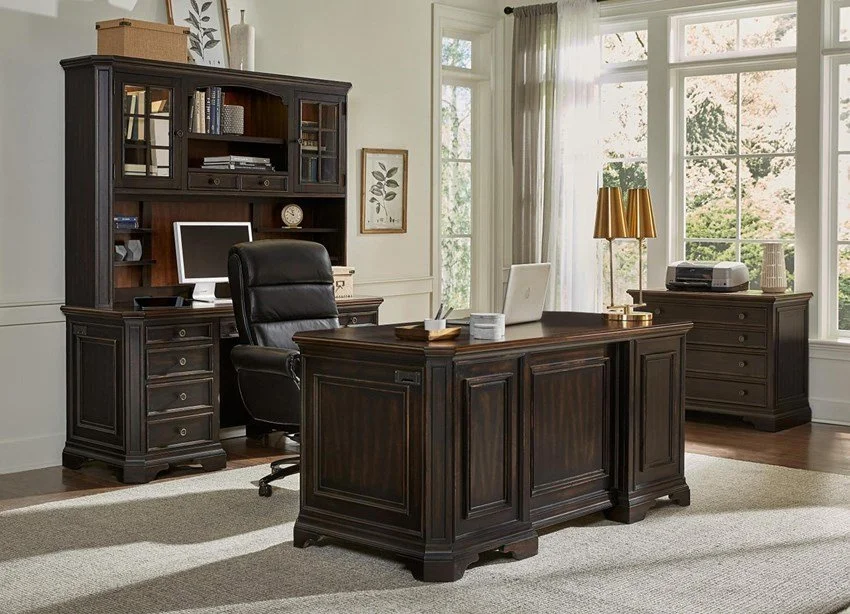 Home office with dark wooden desk, bookcase, and sideboard, black executive chair, computer, lamp, and decor, with large windows and natural light.