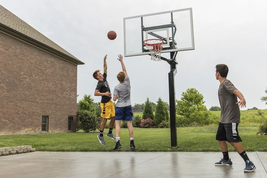 Three boys playing basketball outside near a house, with one jumping to shoot or block the ball at the hoop, another trying to block, and the third watching.