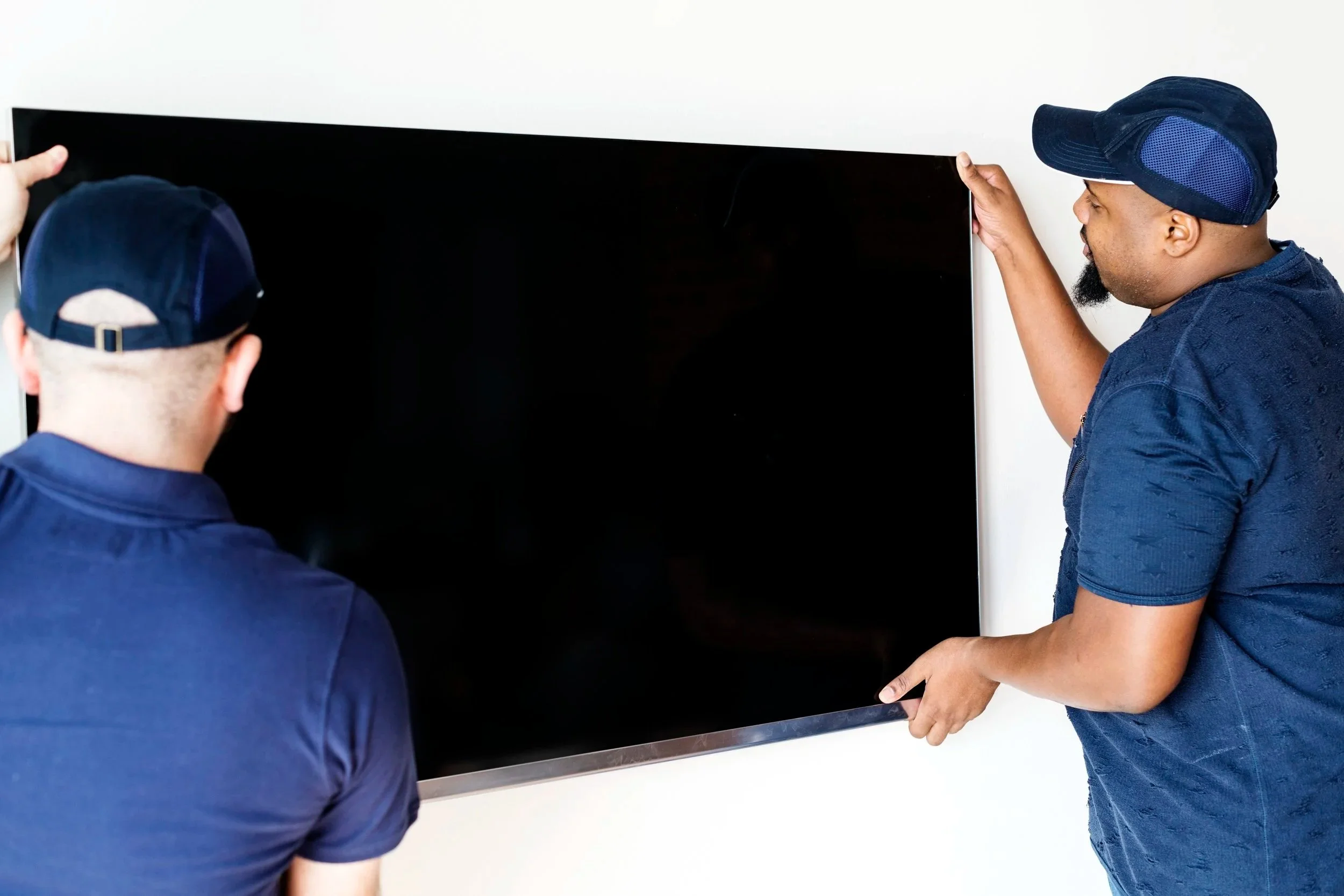 Two men, one wearing a baseball cap and the other a navy cap, are installing or removing a large flat screen television from a white wall.