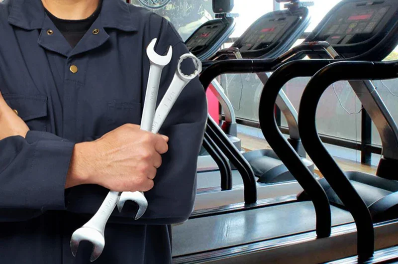 A person wearing a navy uniform holding two wrenches, standing in front of exercise treadmills in a gym.