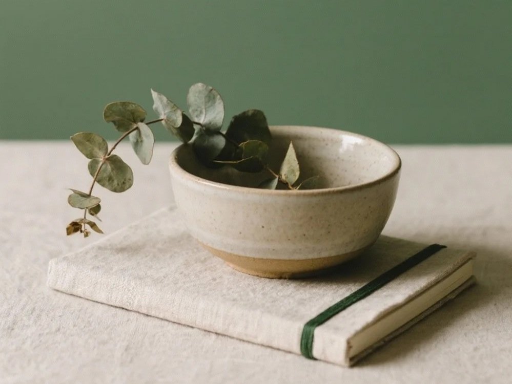 A ceramic bowl containing green eucalyptus leaves, resting on a white cloth napkin with a dark elastic band, placed on a flat surface with a green and beige background.