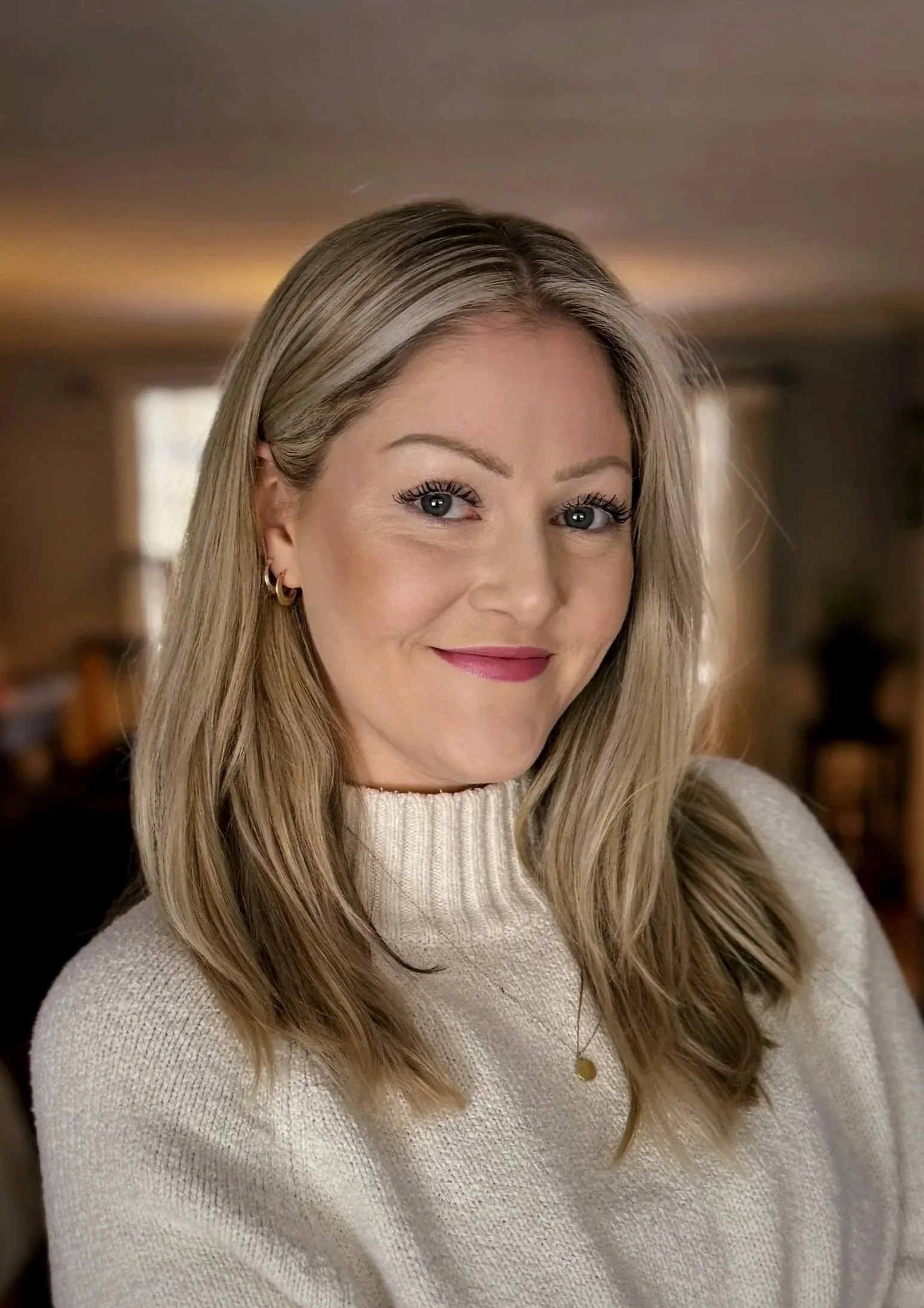 Close-up of a woman with long blonde hair wearing a beige sweater, smiling softly in a warmly lit indoor setting.