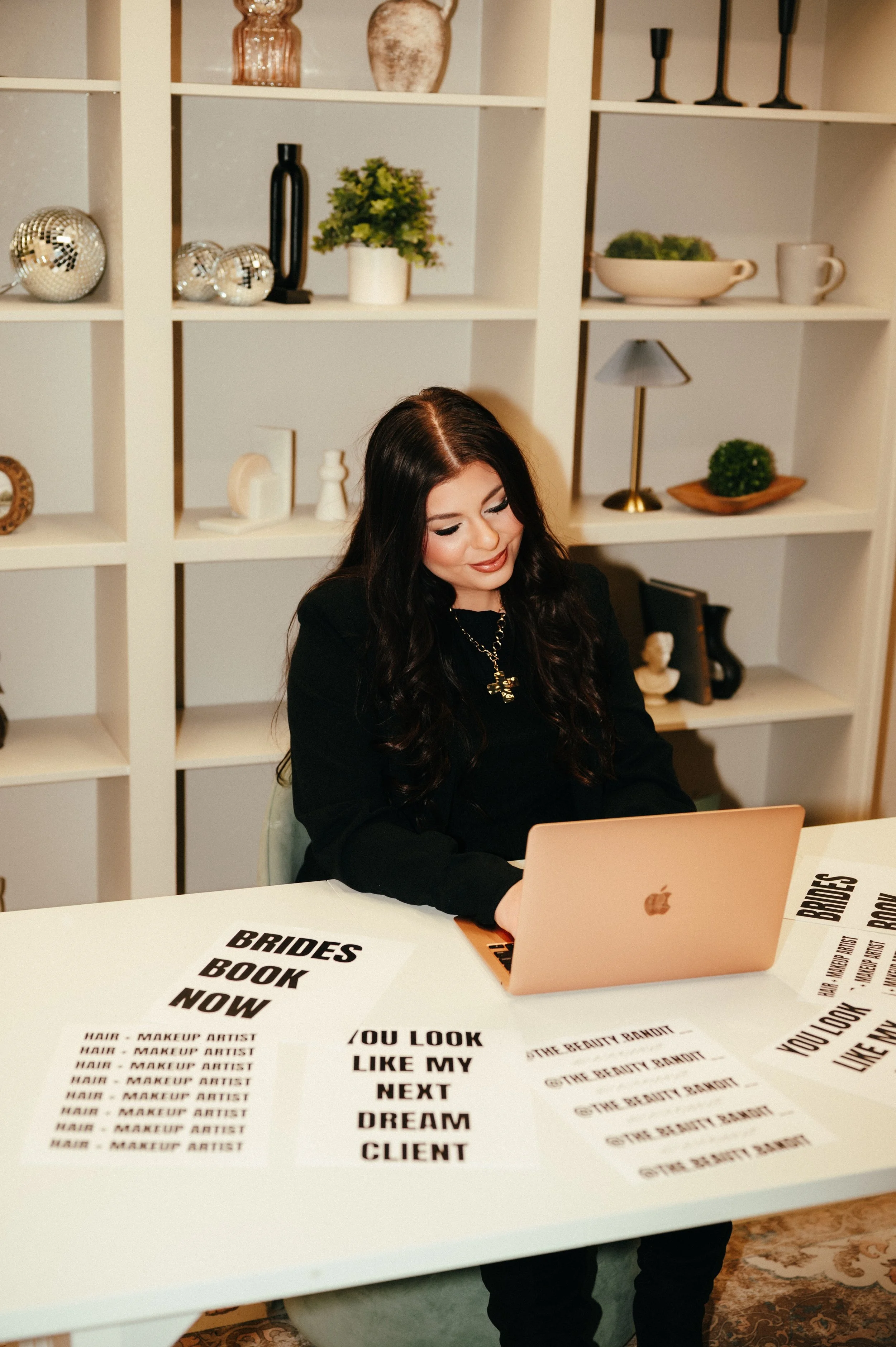 A woman with long dark hair is sitting at a white table with printed signs spread out in front of her, working on a laptop in a room with a white bookshelf behind her.
