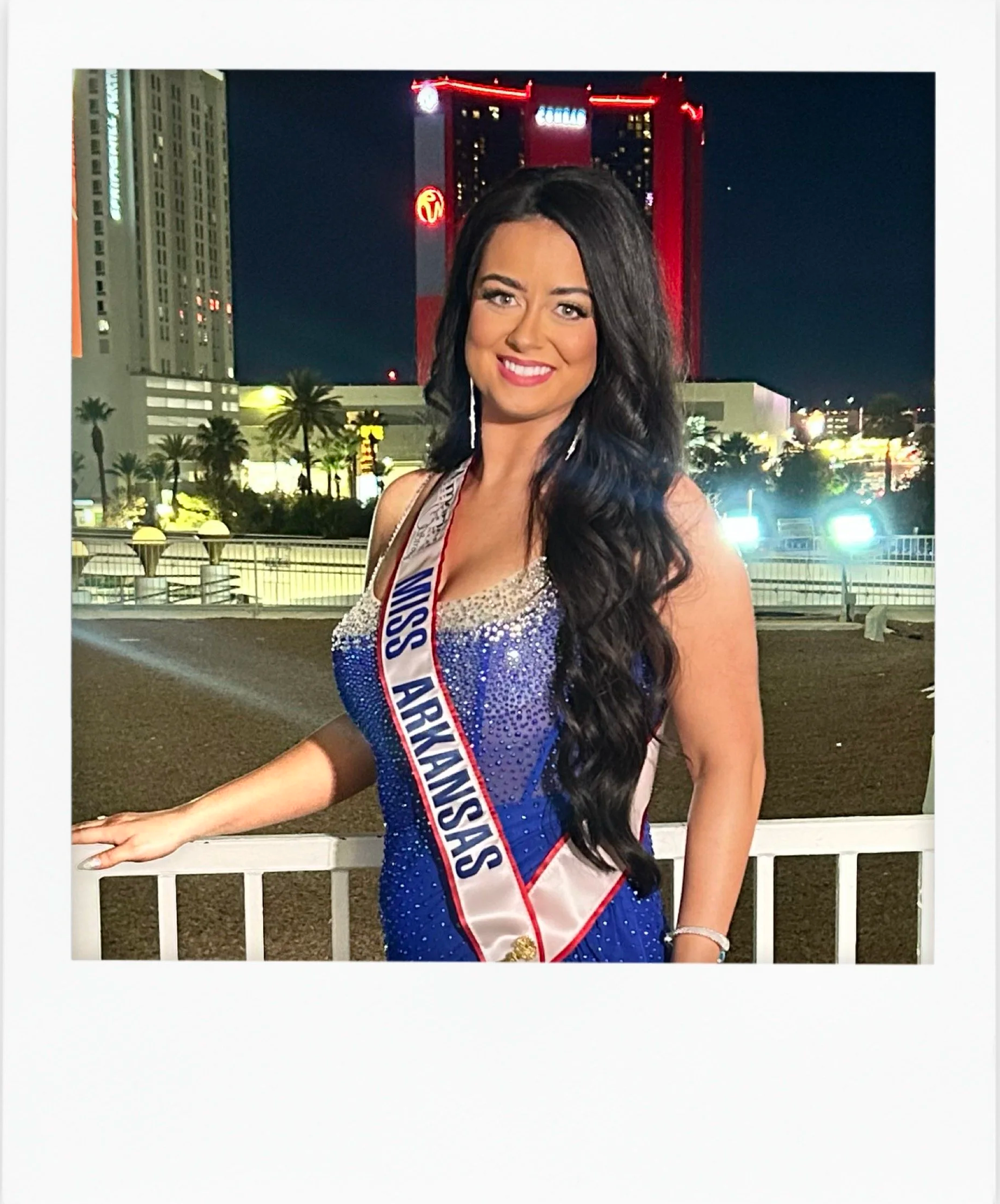 Miss Arkansas wearing a blue evening gown and sash, posing at night with city buildings and bright lights in the background.