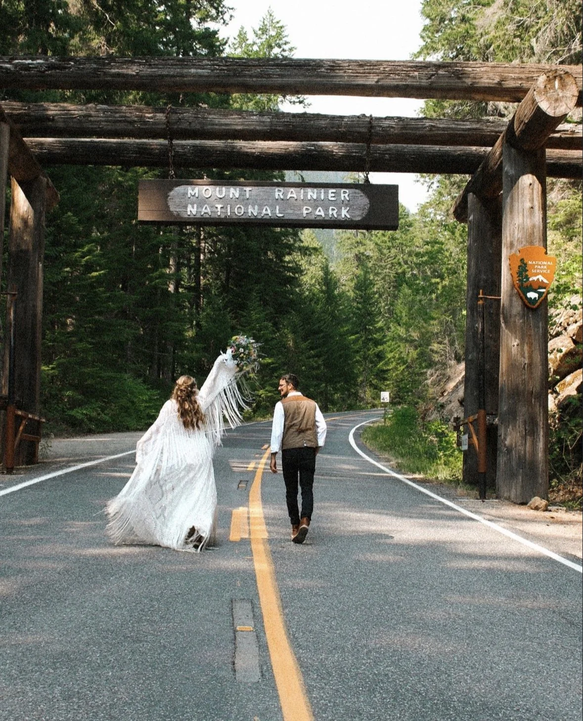 A couple walking on a road through the entrance of Mount Rainier National Park, surrounded by trees, with a wedding veil and flowers.