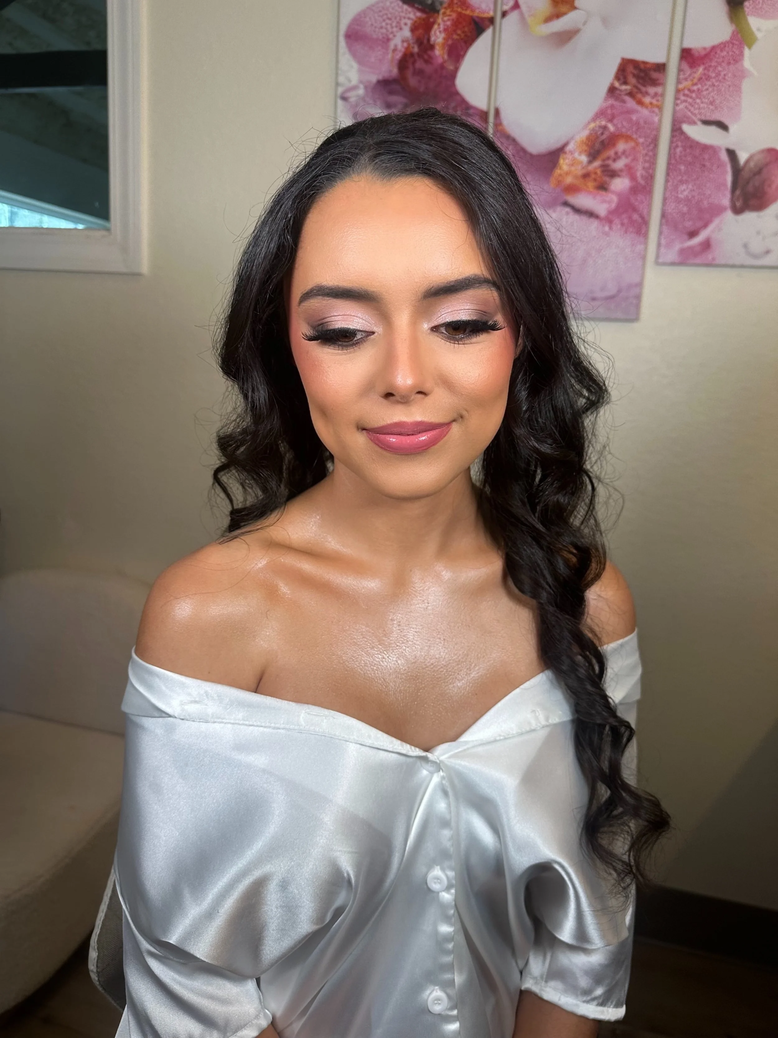 A woman with long dark hair styled in loose curls, wearing makeup including pink lipstick, eyeshadow, and false eyelashes, smiling softly with her eyes closed. She is dressed in an off-the-shoulder white satin blouse, revealing her shoulders and coll