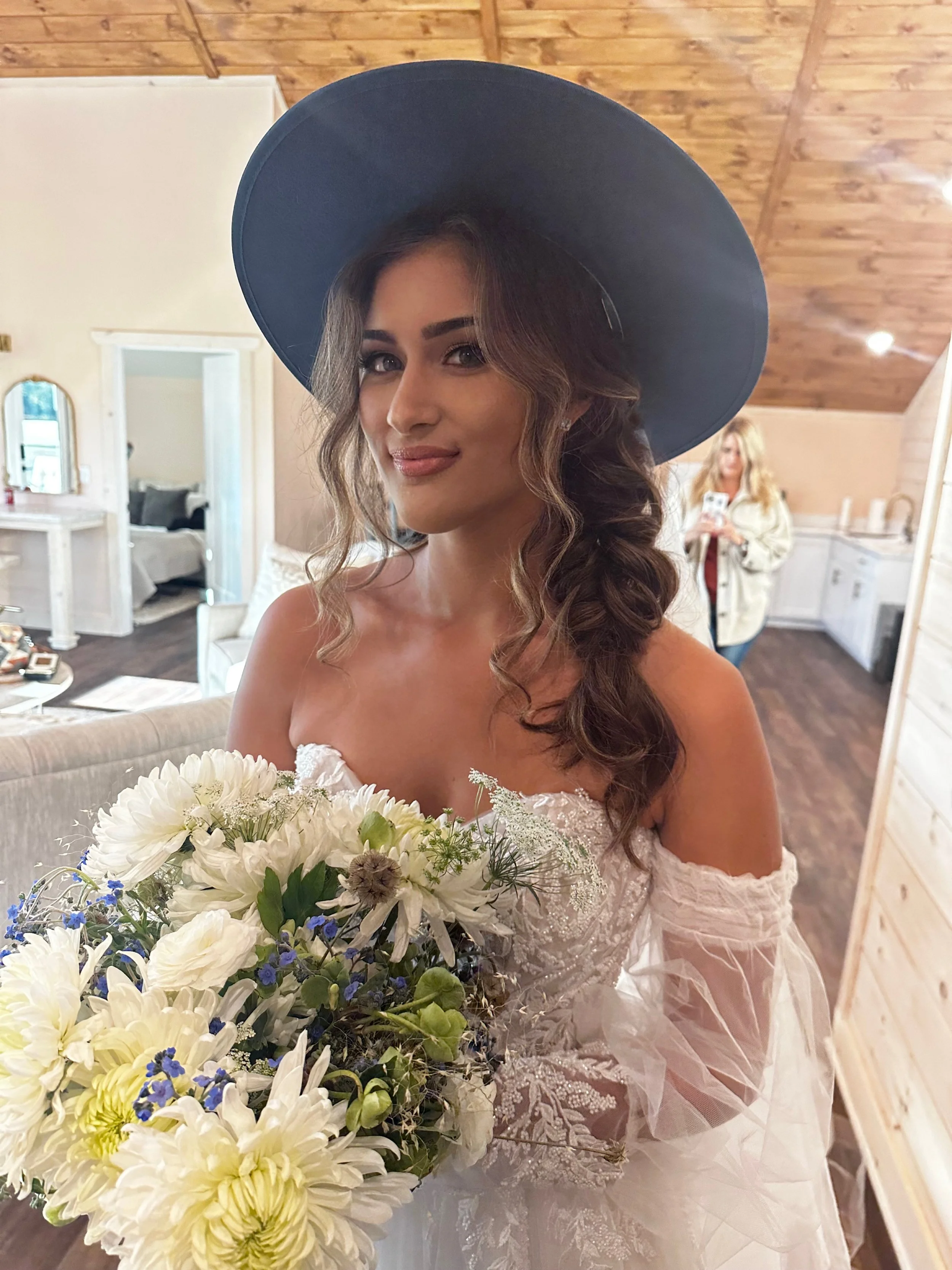 A woman wearing a large black hat and a white off-the-shoulder lace wedding dress, holding a bouquet of white and purple flowers, standing inside a room with wooden ceiling and walls.