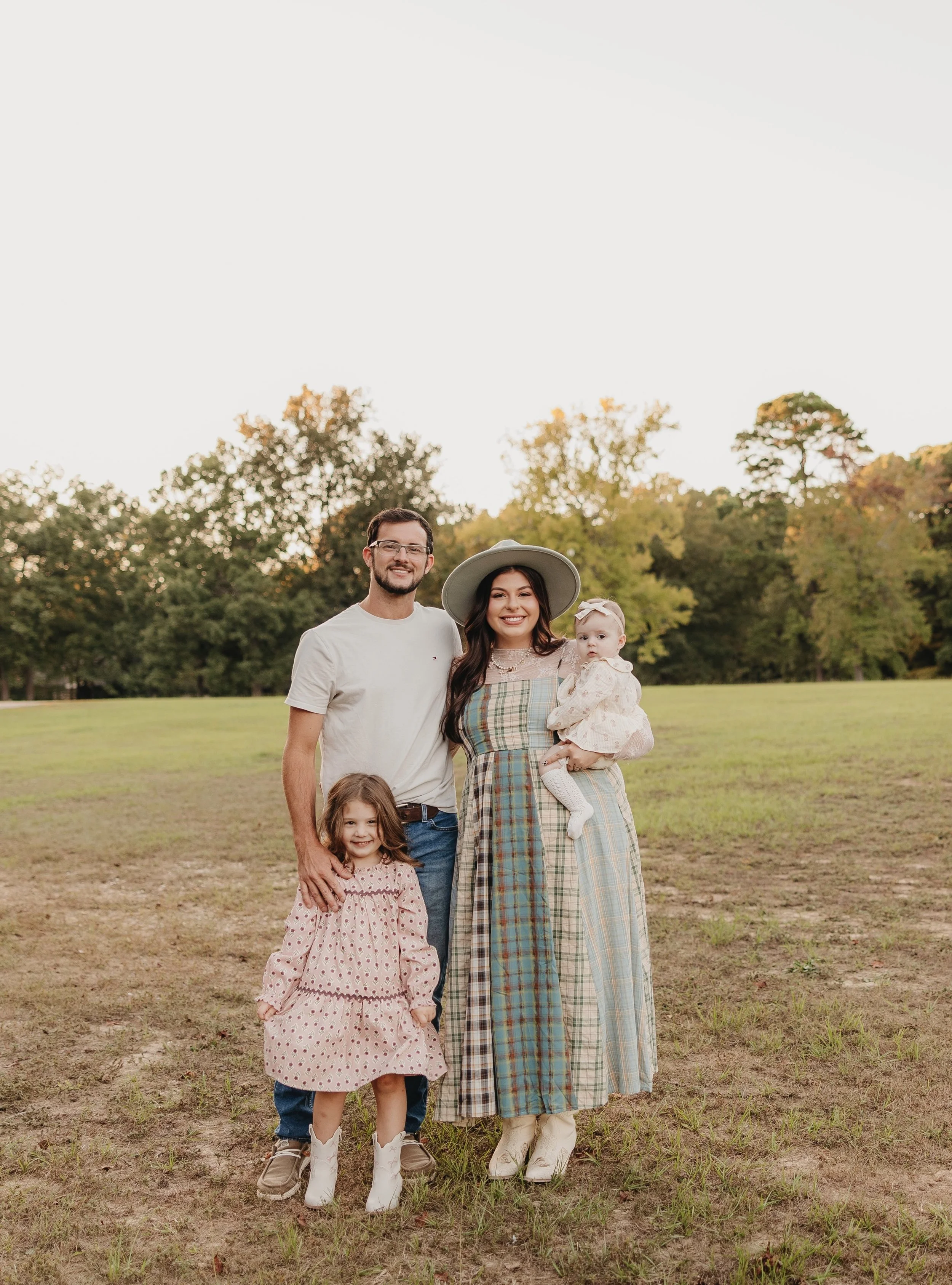 A family of four standing outdoors on a grassy field with trees in the background during sunset. The father wears glasses and a white t-shirt, the mother wears a wide-brimmed hat and a multicolored plaid dress, the young girl wears a pink dress with polka dots, and the baby girl wears a light-colored dress with a bow and white socks.