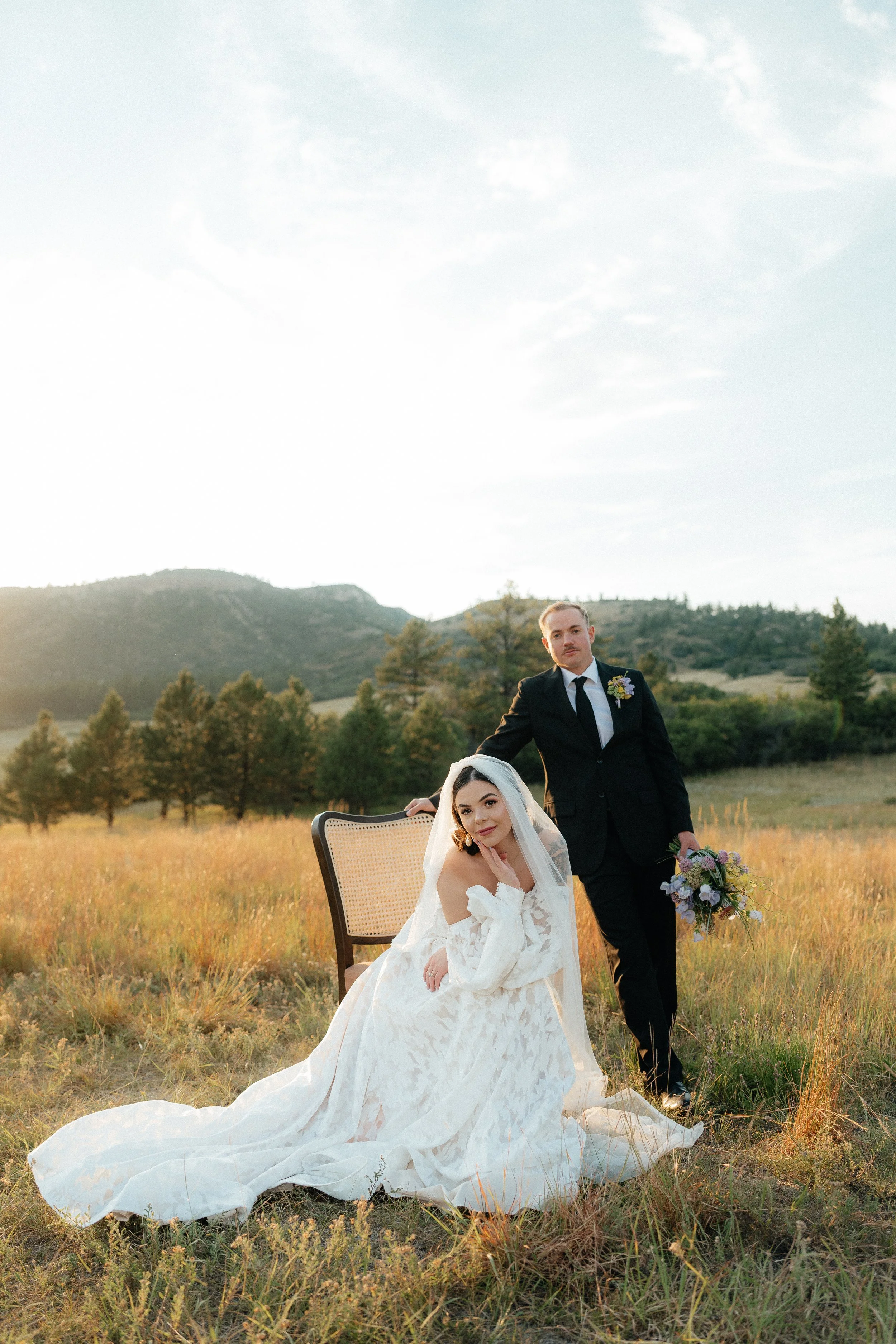 A bride and groom posing in a field with mountains in the background during sunset. The bride is sitting on the ground in a white wedding dress, and the groom is standing next to her in a black suit holding a floral bouquet.