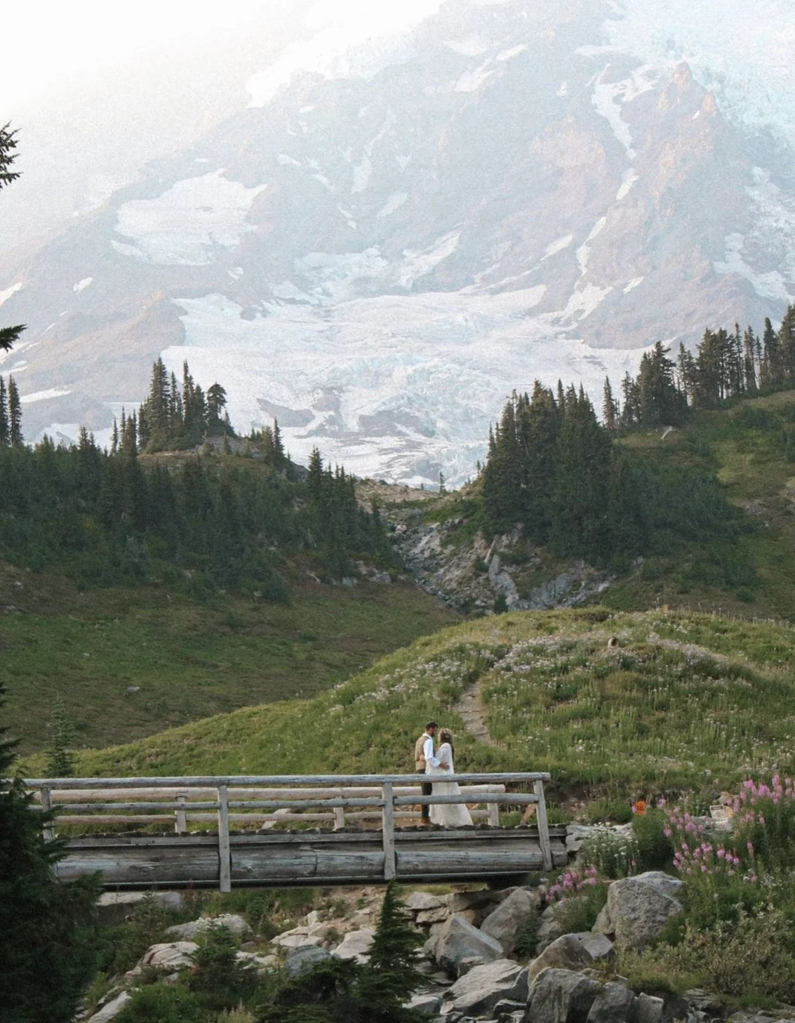 A couple standing on a wooden bridge in a mountainous landscape with snow-capped peaks in the background and wildflowers on the hillside.