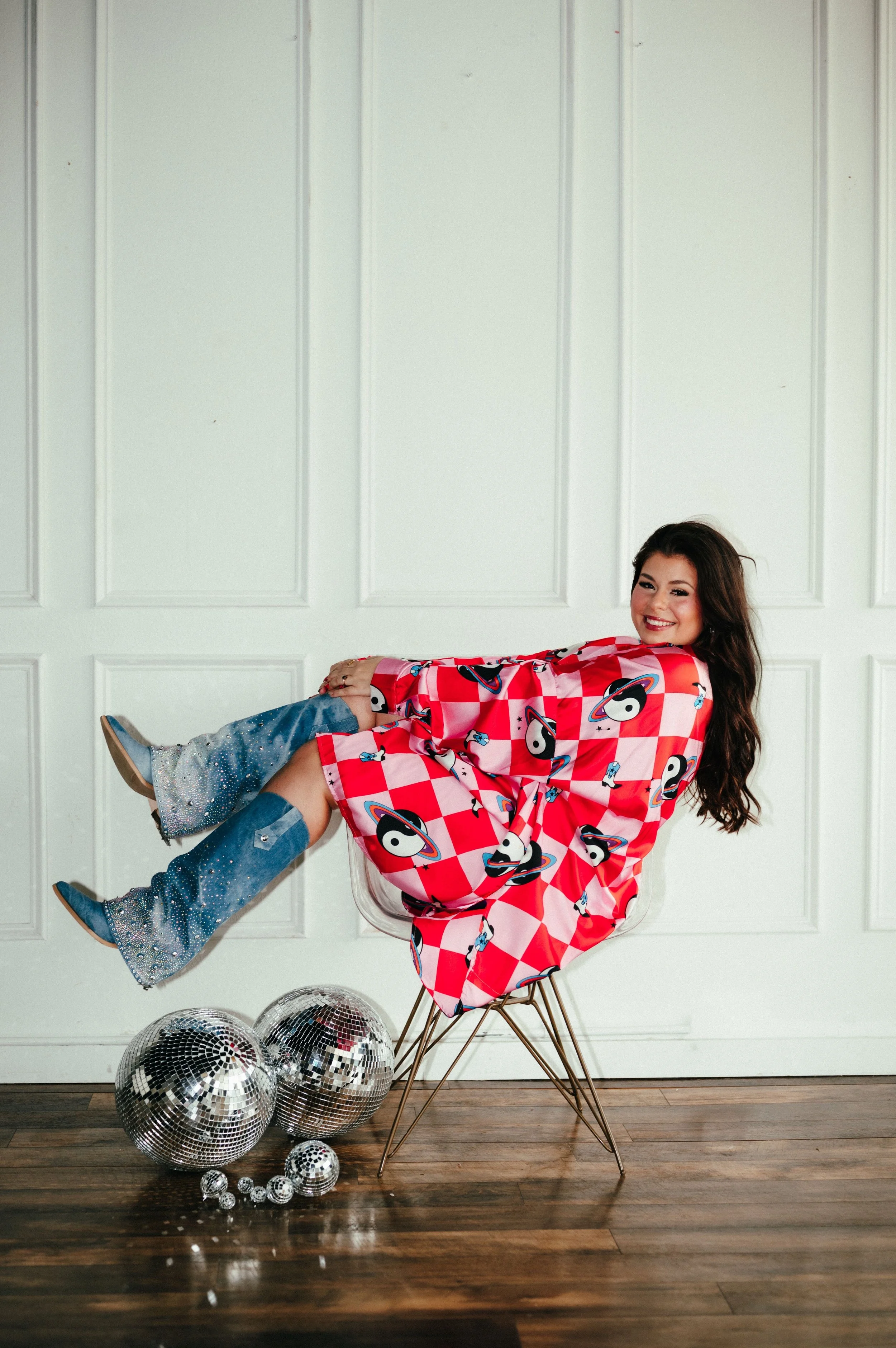 A woman sitting on a chair, wearing a red checkered robe with yin-yang and planet designs, blue cowboy boots with rhinestones, smiling at the camera, surrounded by disco balls on a wooden floor in front of a white paneled wall.