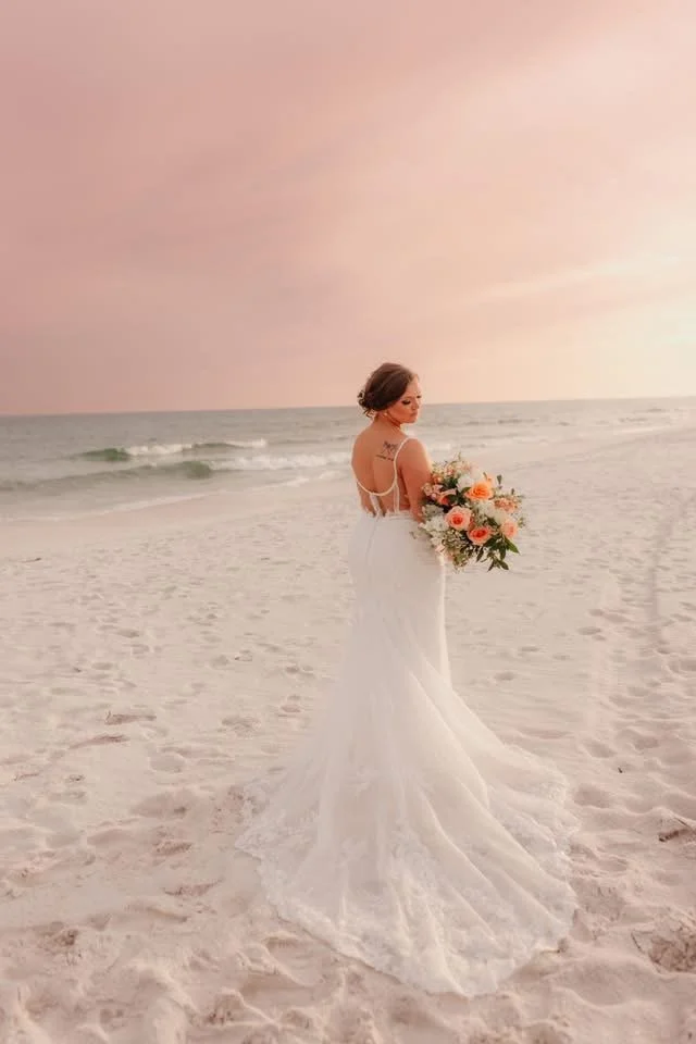 A bride in a white wedding dress holding a bouquet of flowers on a beach at sunset.