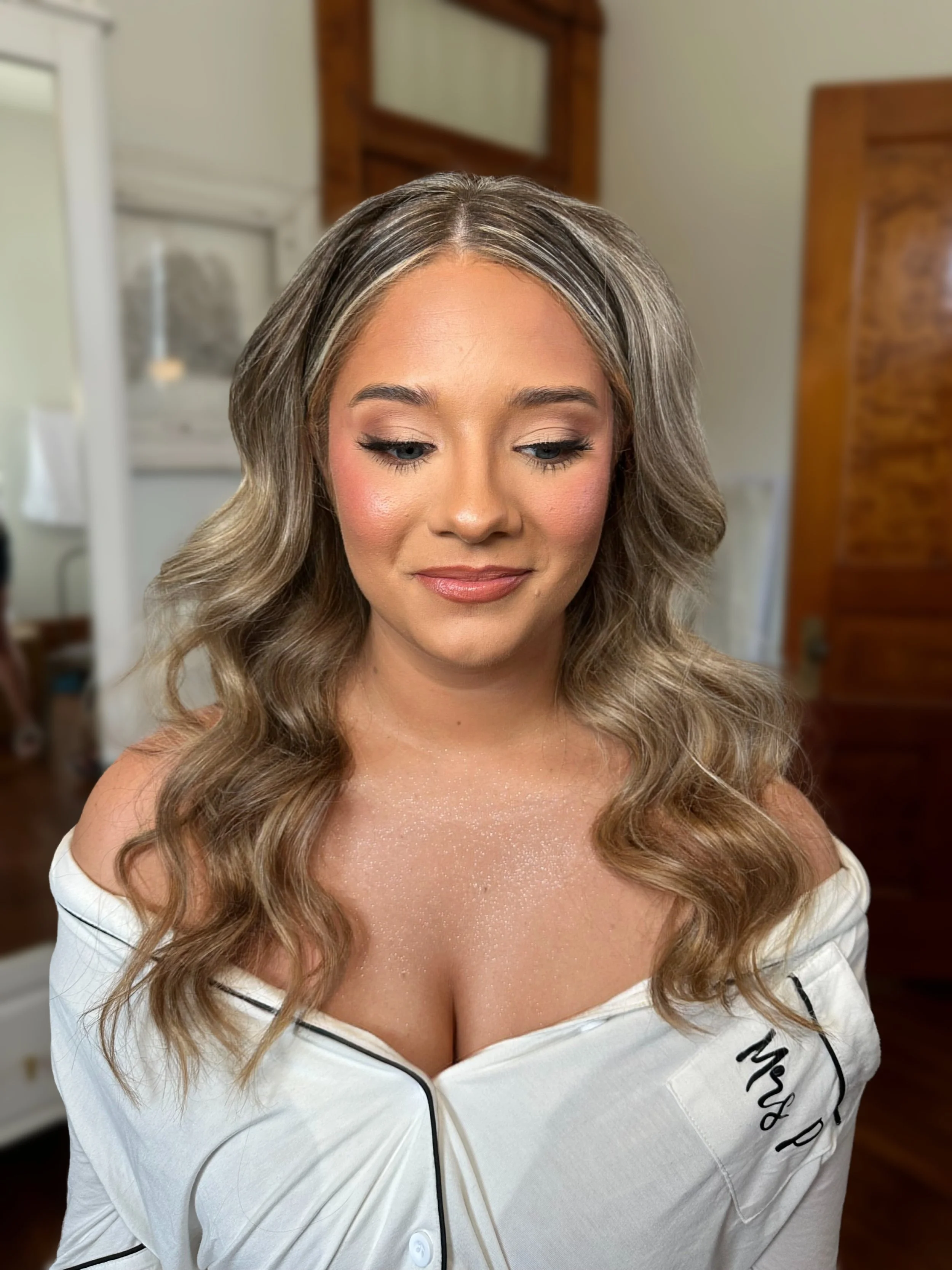 A woman with wavy blonde hair, wearing makeup, and dressed in a white off-shoulder top with black piping and the words 'Mrs P.' embroidered on the shoulder, standing indoors in a room with wooden furniture.