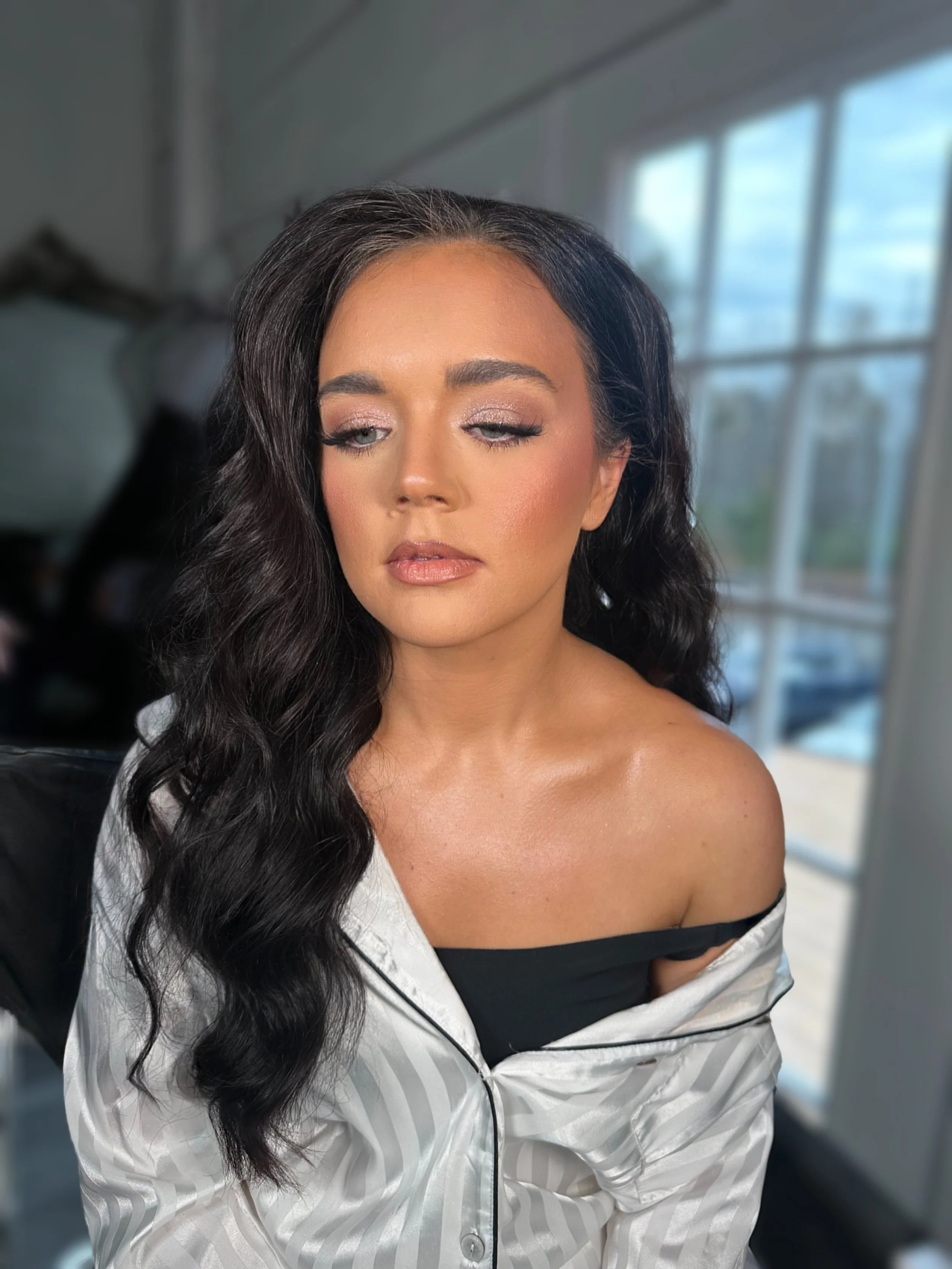 A woman with wavy black hair, wearing makeup, partially dressed in a black top and a satin pajama top, in a room with large windows showing a cloudy sky.