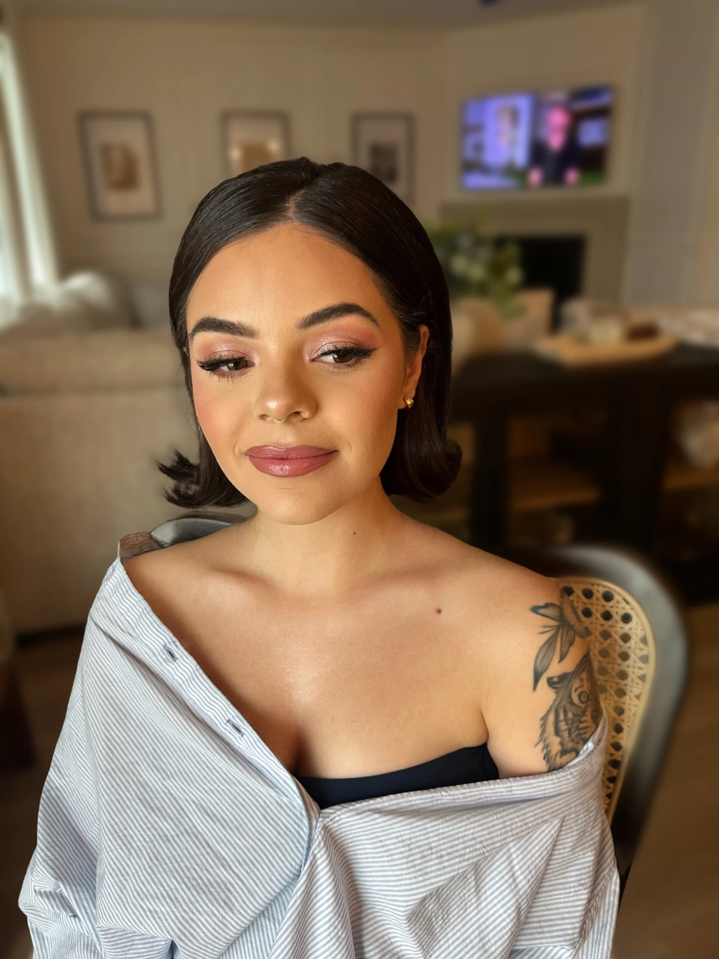 A young woman with shoulder-length dark hair, makeup, and jewelry, sitting in a home interior with a blurred background, wearing a striped shirt falling off one shoulder to reveal a tattoo of a tiger and leaves on her upper arm.