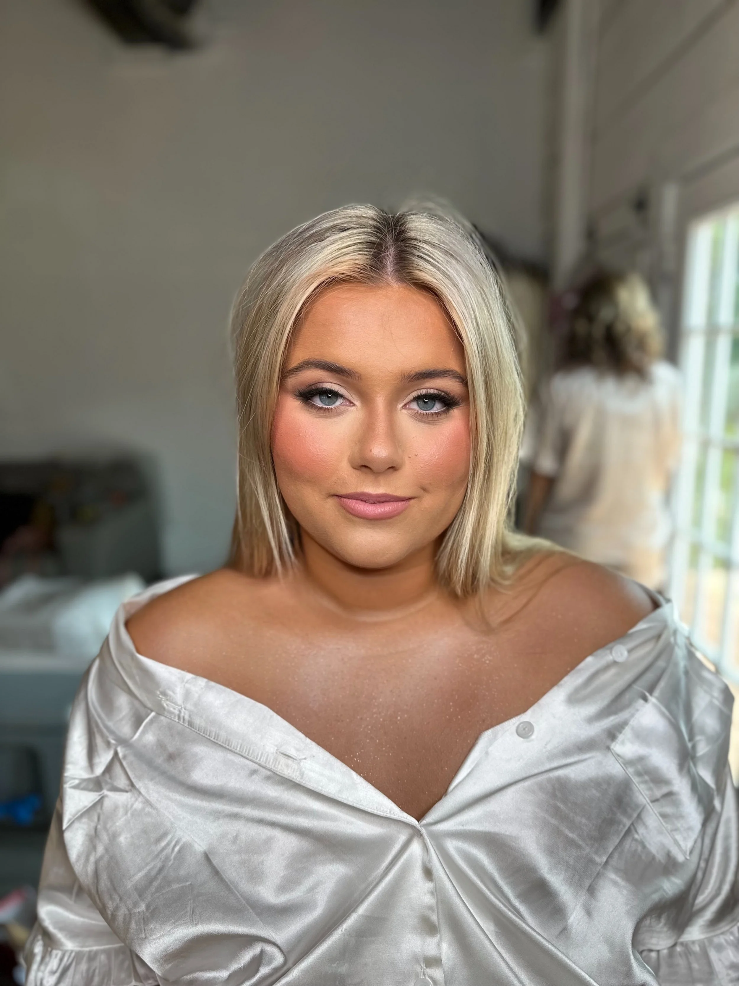 A young woman with blonde hair and blue eyes, wearing a partially unbuttoned white silky top, stands in a well-lit room with a person in the background.