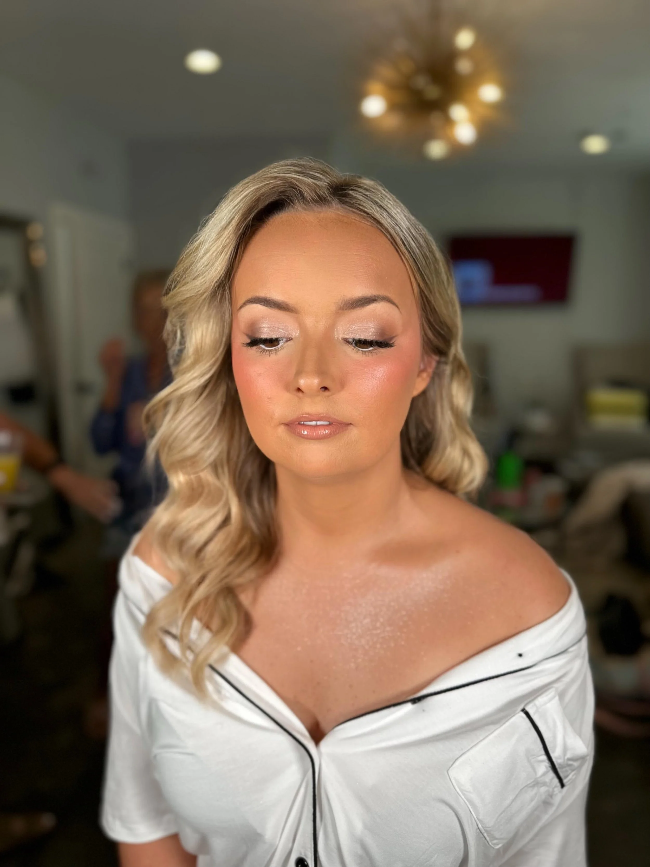 A woman with blonde wavy hair styled in loose curls and makeup, wearing an off-the-shoulder white robe with black piping, standing in a room with a blurred background.