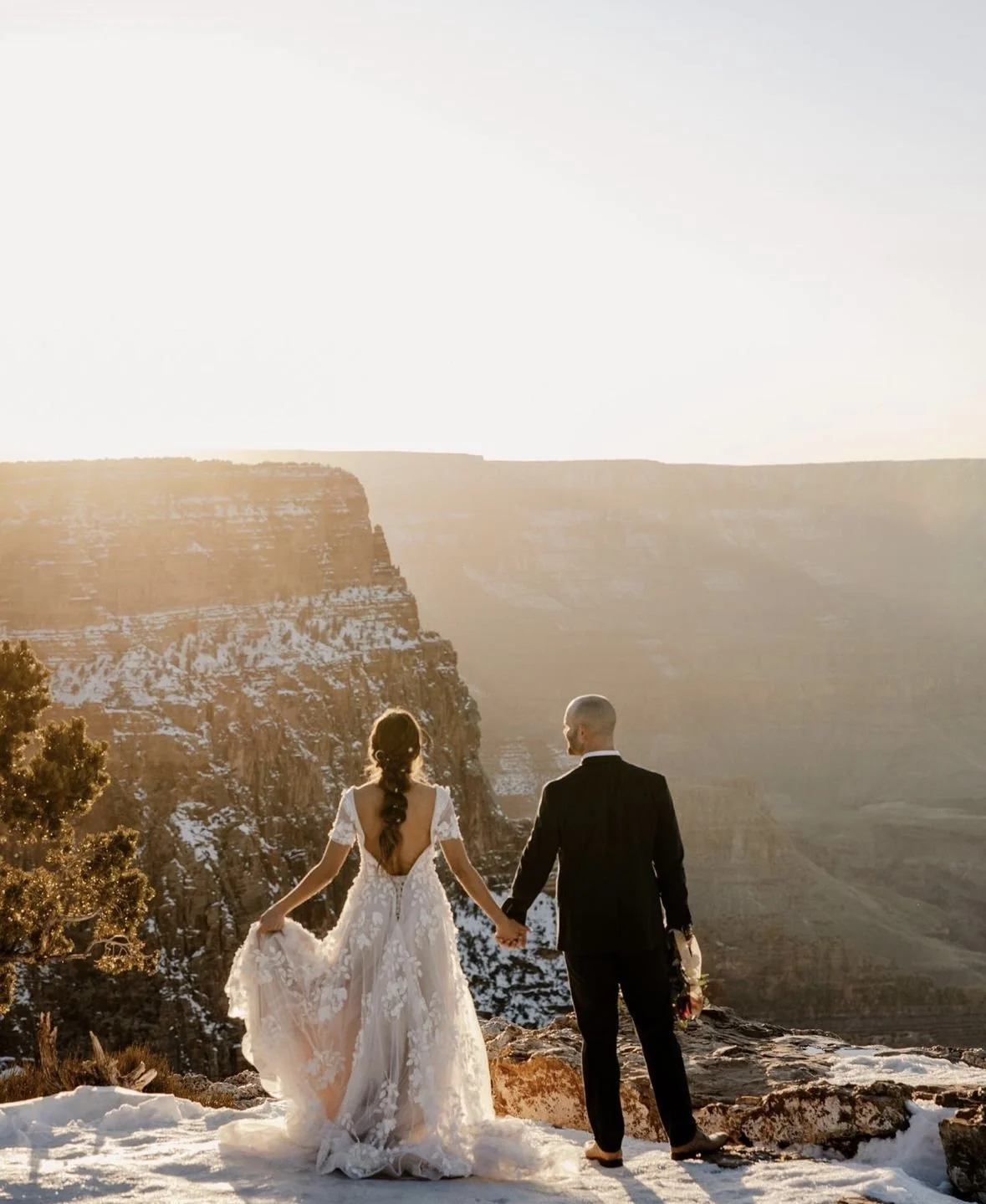 A bride and groom holding hands on a snowy cliff, with the Grand Canyon in the background at sunset.