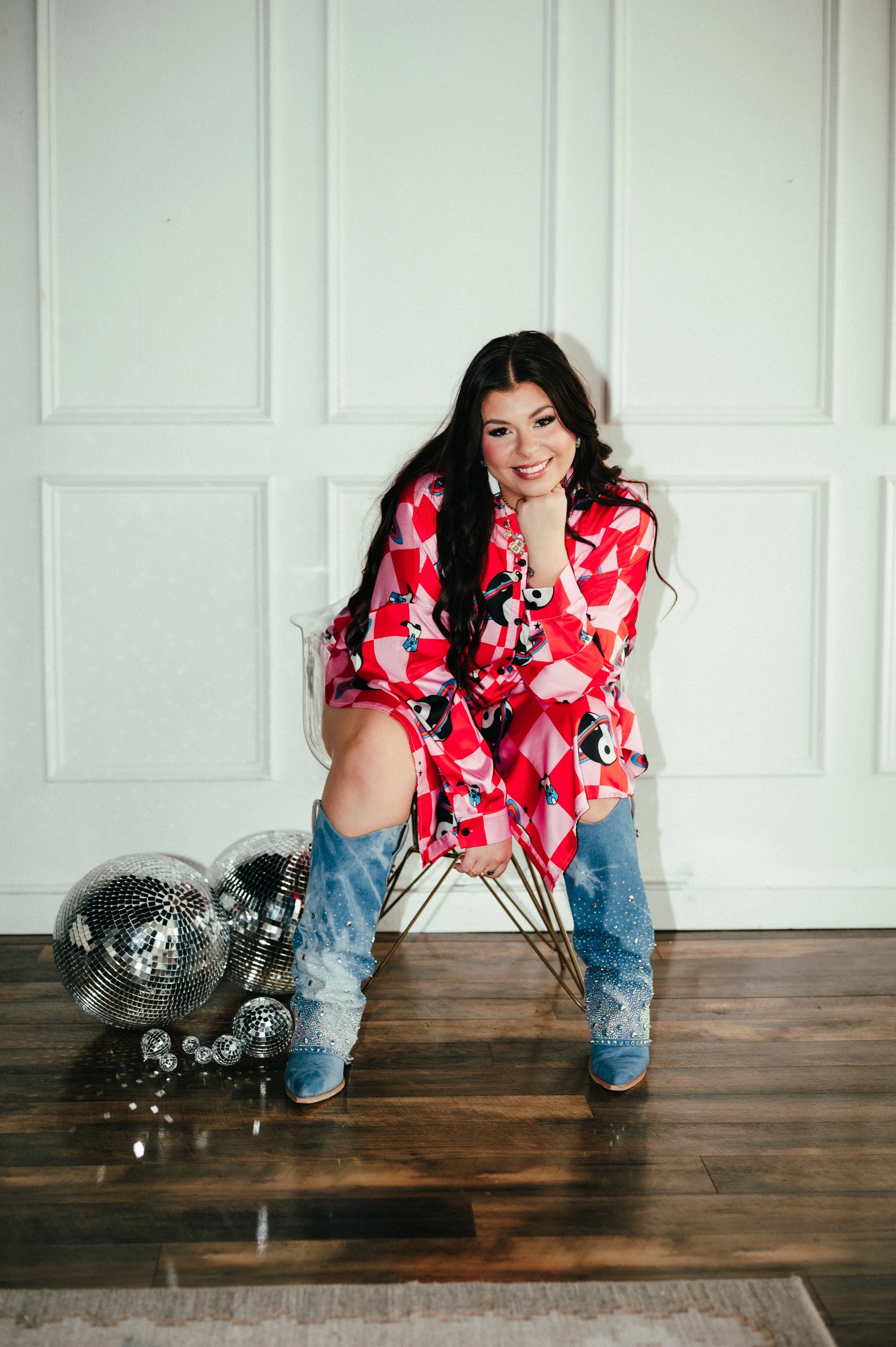 A woman with long dark hair, wearing a colorful pink patterned dress and light blue embellished boots, sitting on a gold wire chair in front of a white paneled wall. Disco balls are on the floor next to her.