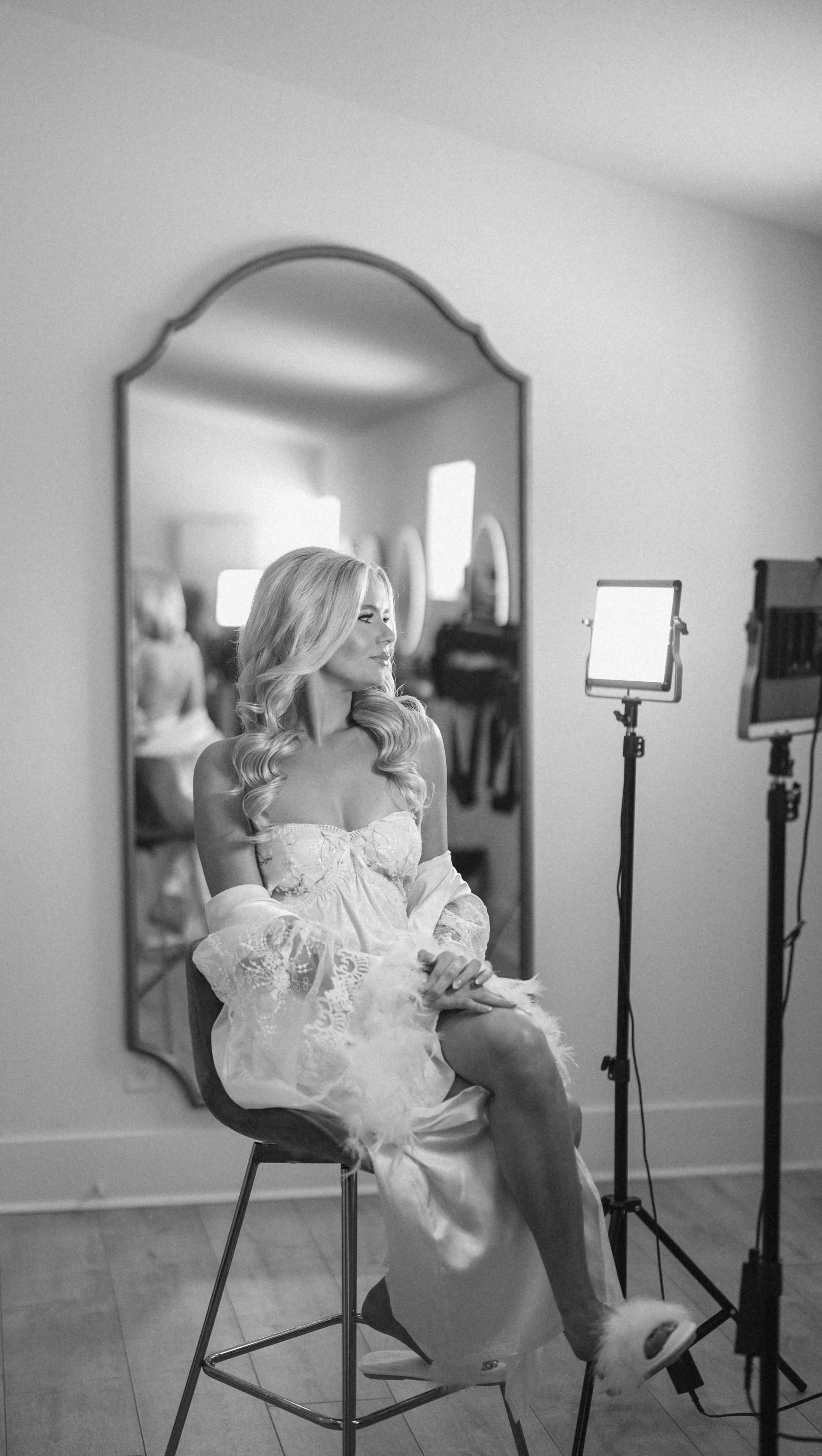 A woman with long blonde hair in a white satin gown with lace and feather details sits on a chair, looking to her right, during a photo shoot in a room with a mirror and studio lights.