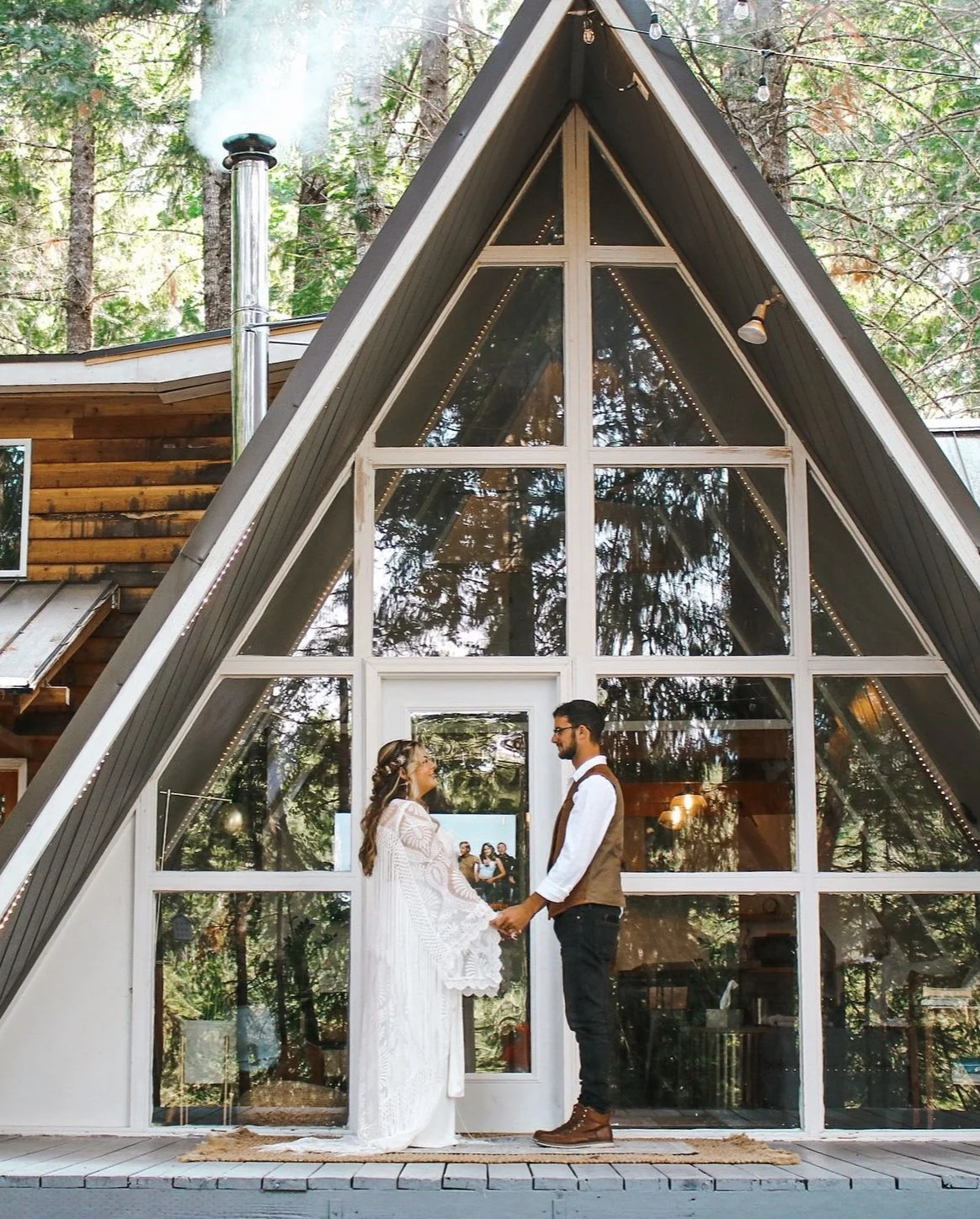 A couple in wedding attire standing in front of a modern A-frame glass house, holding hands and smiling at each other.