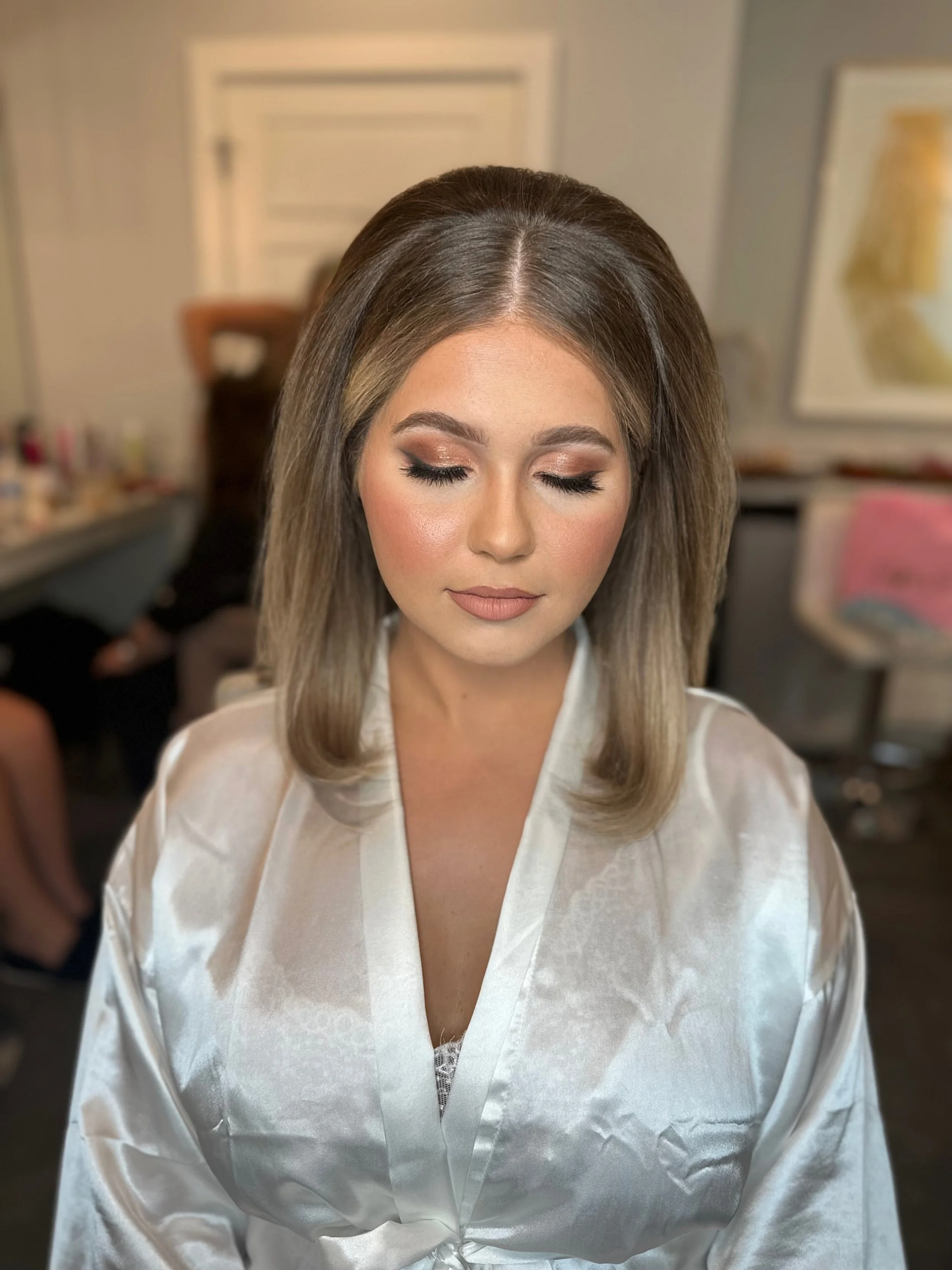 A woman with makeup and hair done, wearing a satin robe, sitting with eyes closed in a room.