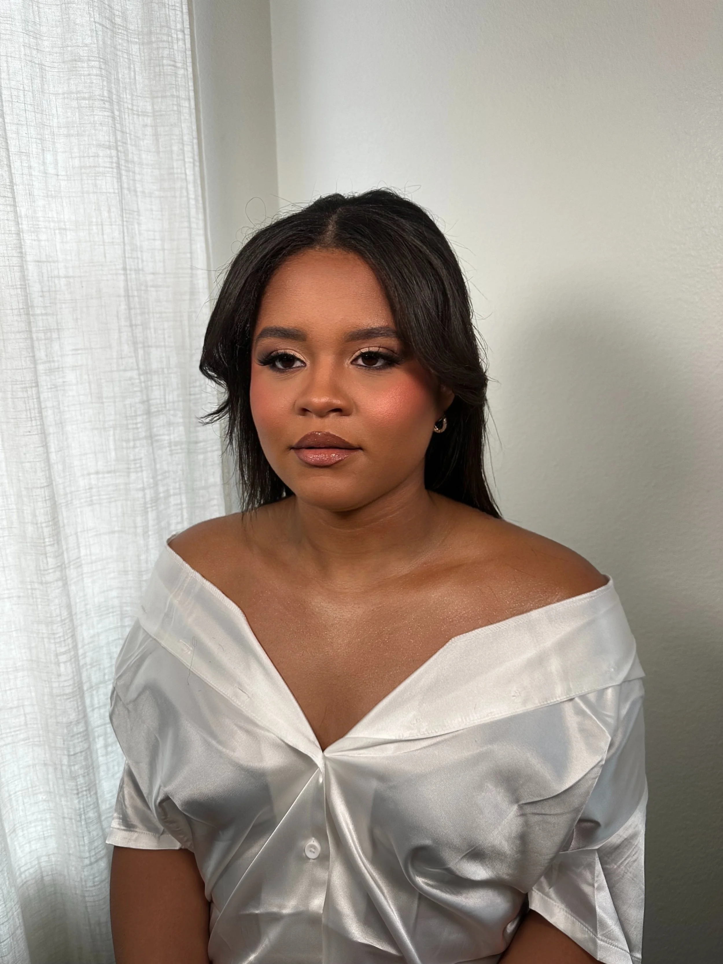 A woman with medium skin tone and dark hair styled in loose curls, wearing makeup with shimmering eyeshadow, eyeliner, blush, and glossy lips. She is dressed in a white satin off-the-shoulder blouse and posed against a light background with sheer whi