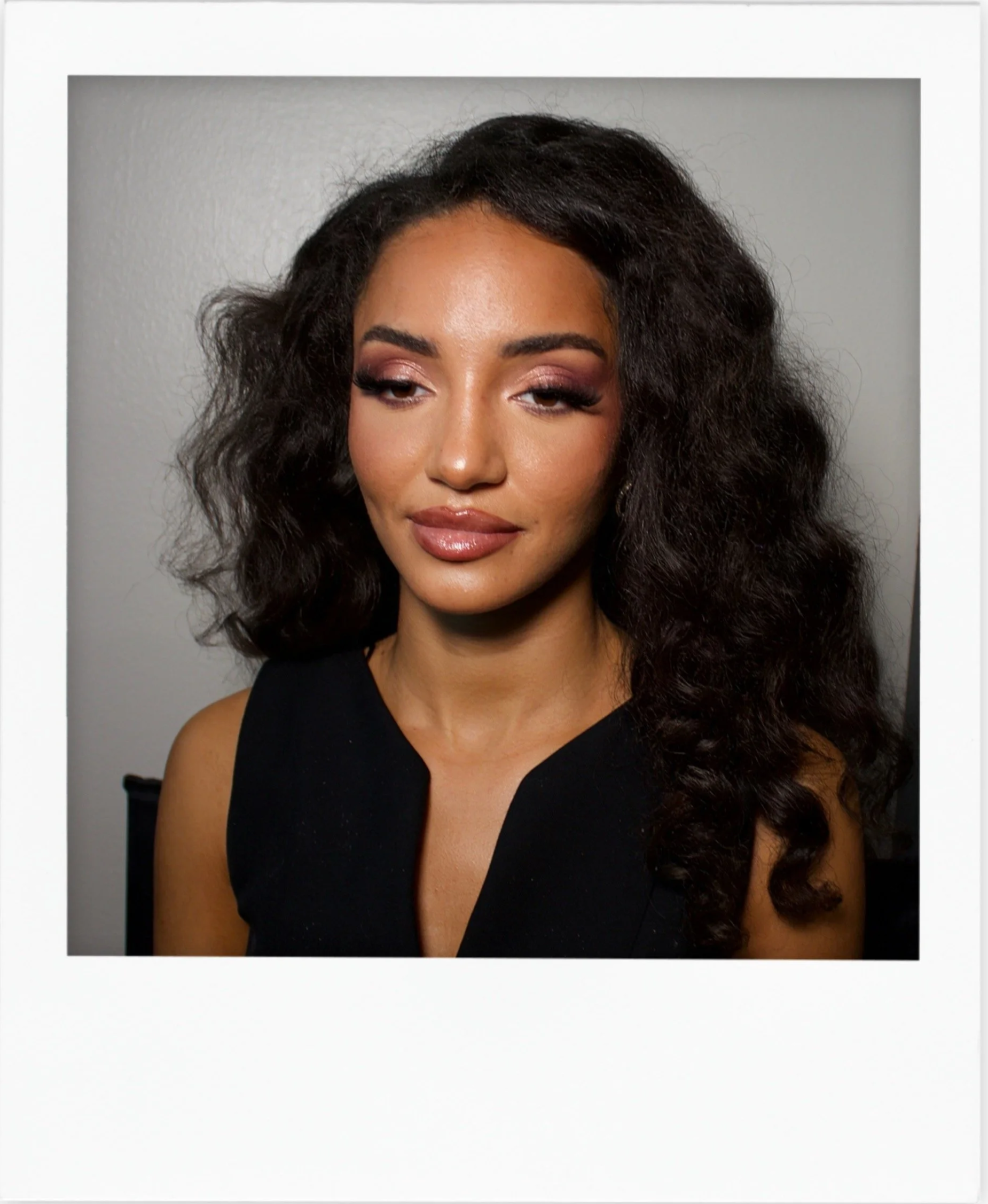 A woman with dark, curly hair and makeup, wearing a sleeveless black top, looking slightly to the side with a neutral expression against a plain gray background.