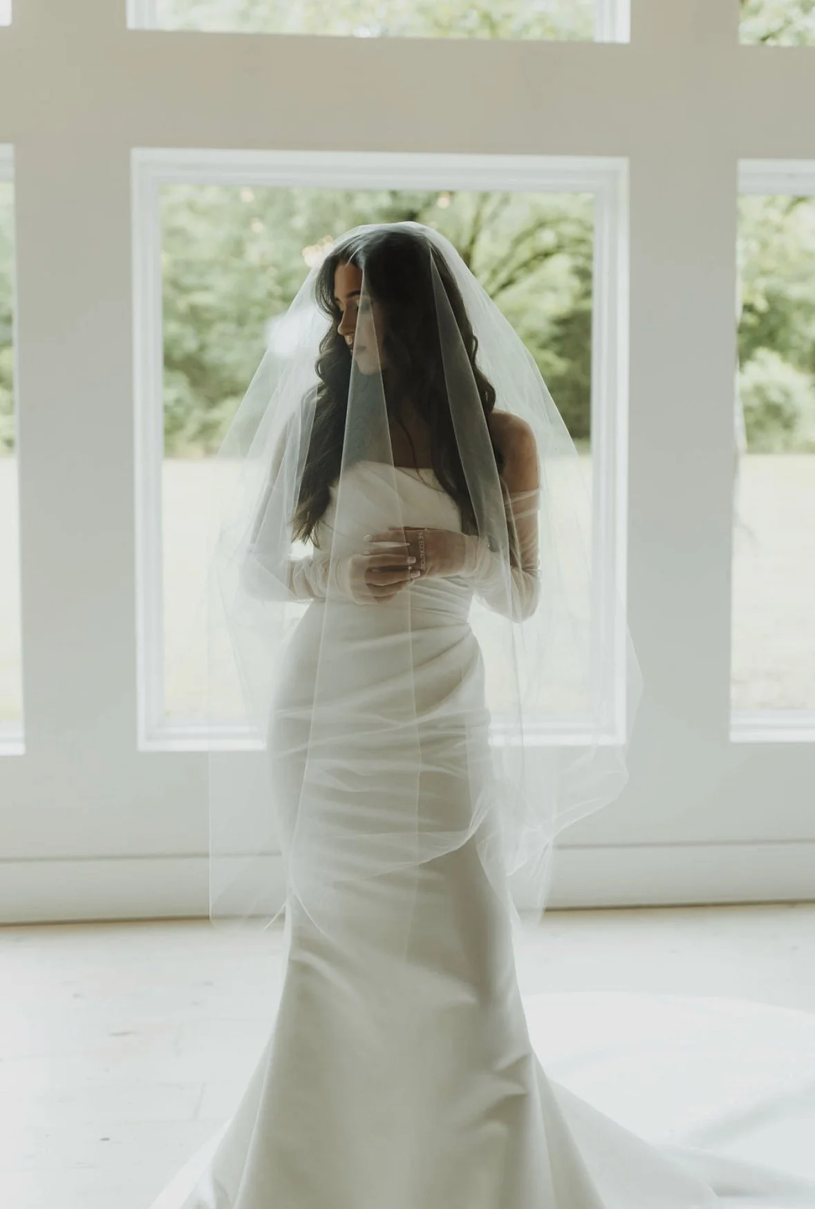 A bride in a white wedding dress and veil standing indoors in front of large windows with trees outside.