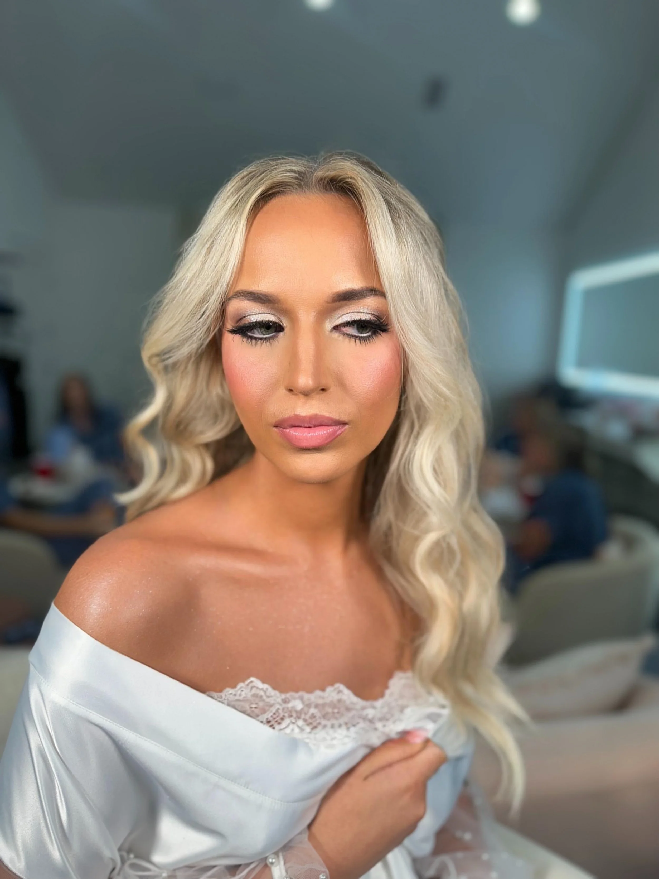 A woman with blonde wavy hair and makeup, wearing a white off-shoulder lace top, sitting in a room with blurred background.