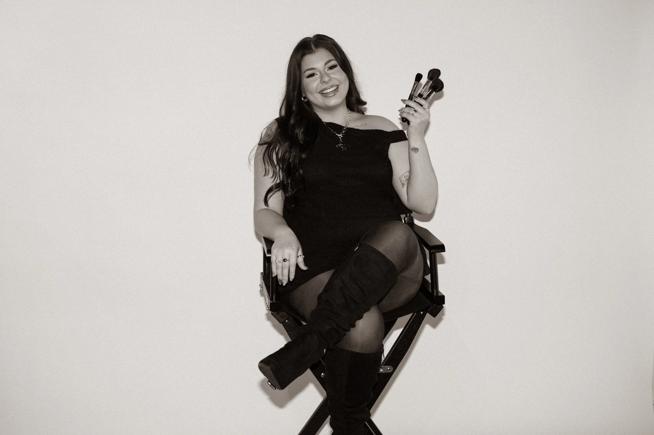A young woman with long dark hair, wearing a black off-the-shoulder dress, sitting on a director's chair, holding makeup brushes, smiling, against a plain light background.