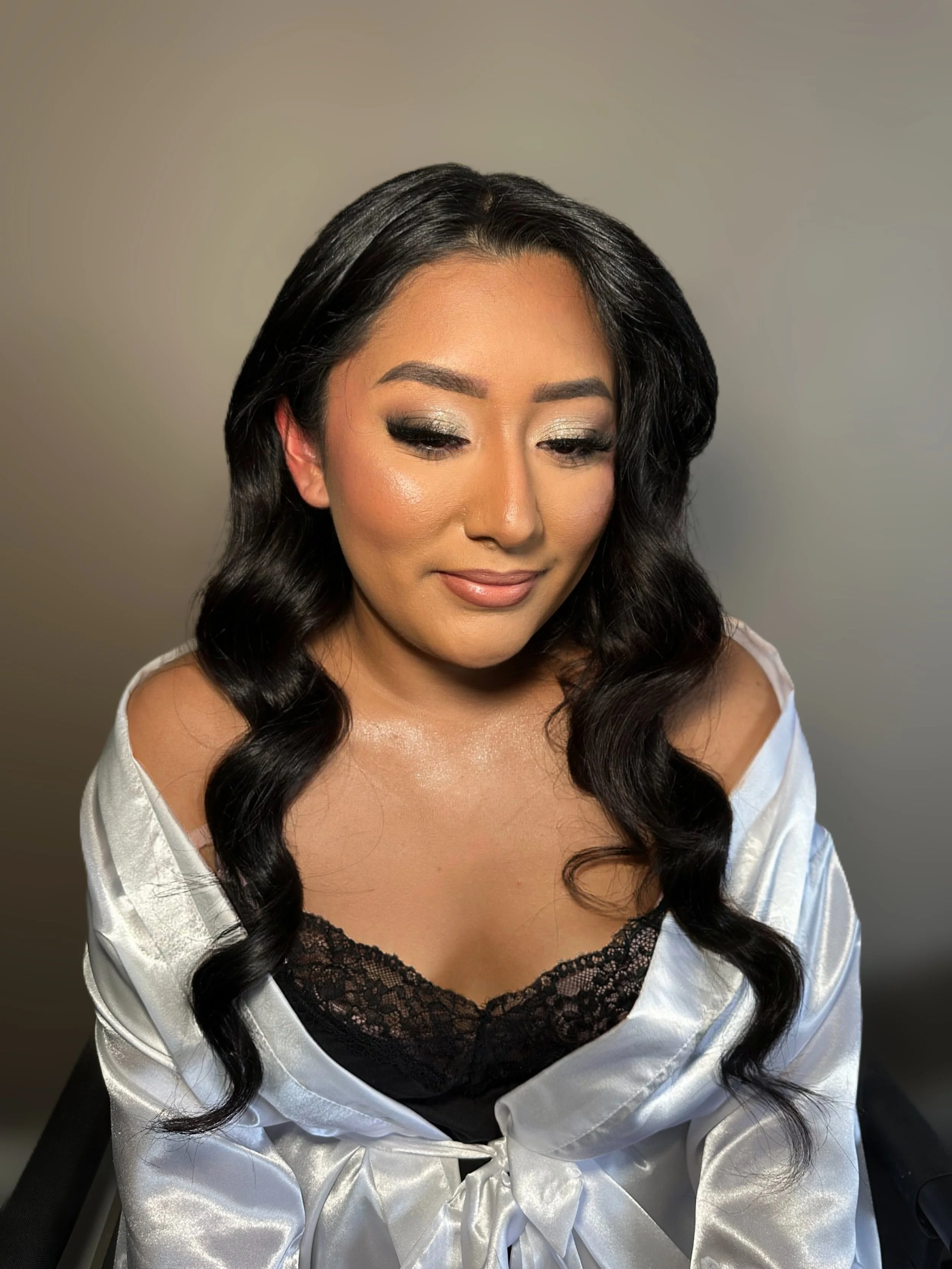 A woman with long black wavy hair, wearing makeup with shimmery eyeshadow and nude lipstick, dressed in a silky satin robe over a black lace top, looking slightly downward with a soft smile.