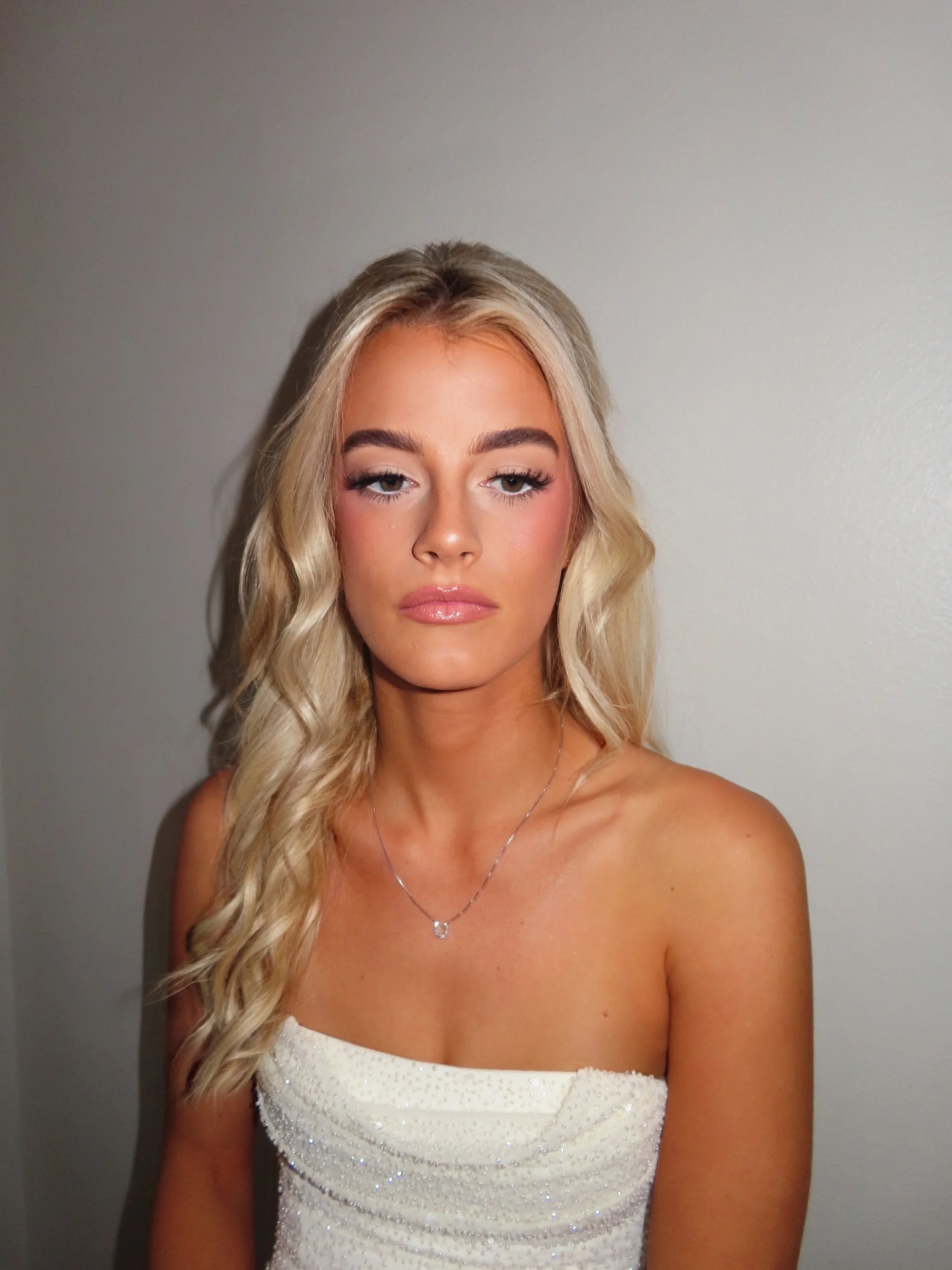 A young woman with blonde wavy hair and fair skin, wearing a strapless white dress with sparkles, silver necklace, and makeup, standing against a plain gray wall.