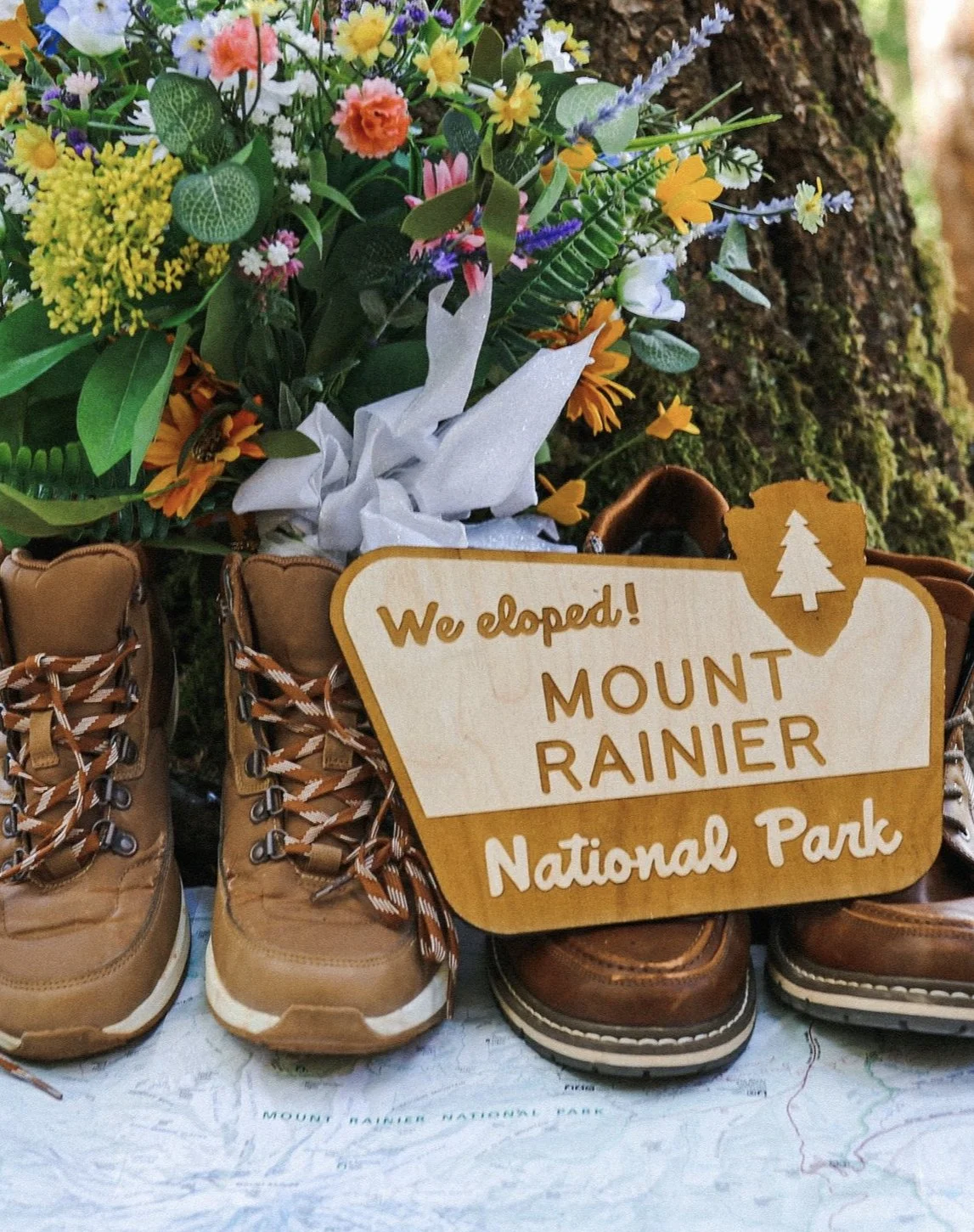 A pair of hiking boots and a wooden sign that says, 'We explored! Mount Rainier National Park,' placed in front of a bouquet of colorful flowers and a tree trunk.