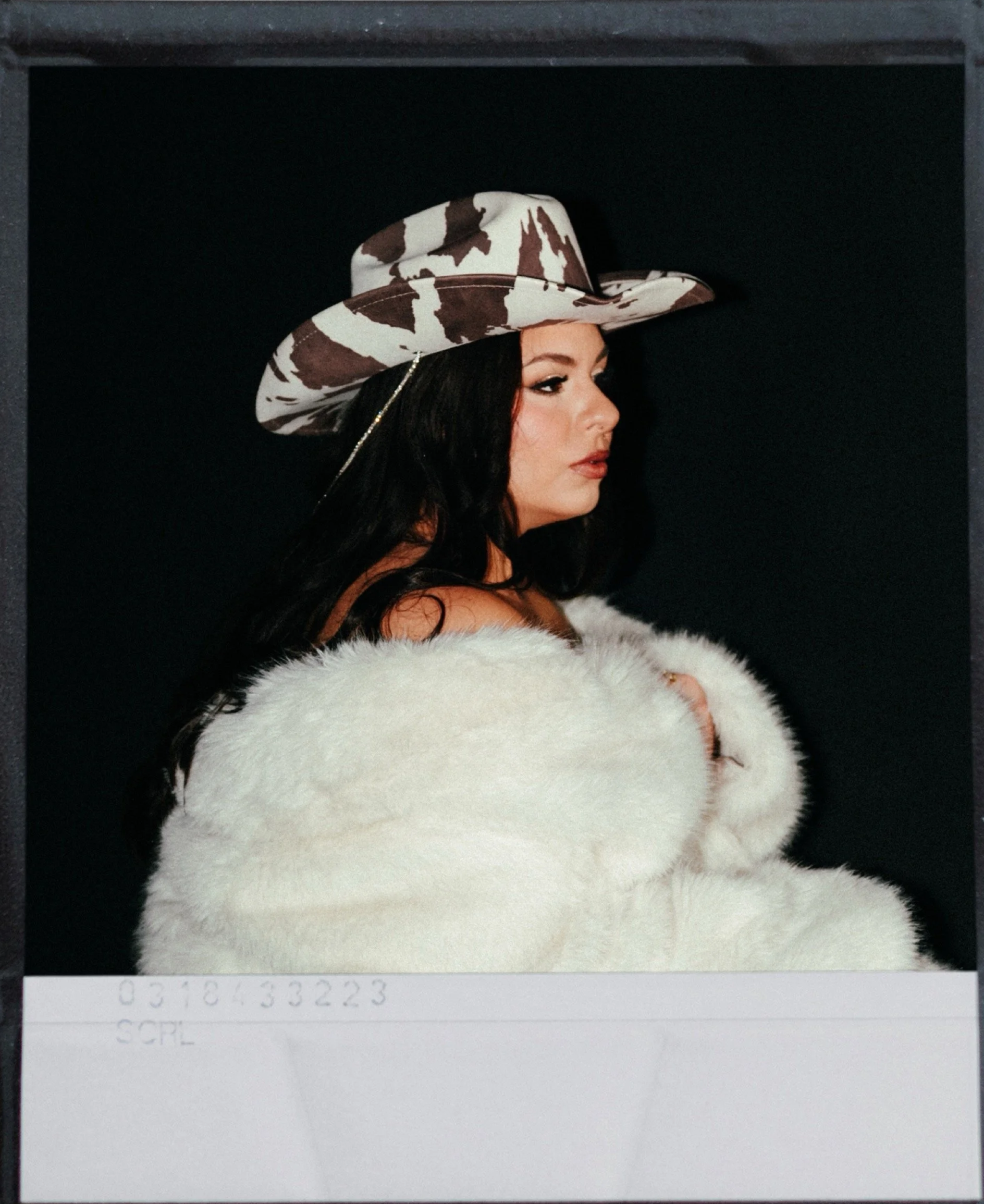 A woman wearing a brown and white patterned wide-brimmed hat and a white fur coat against a black background.