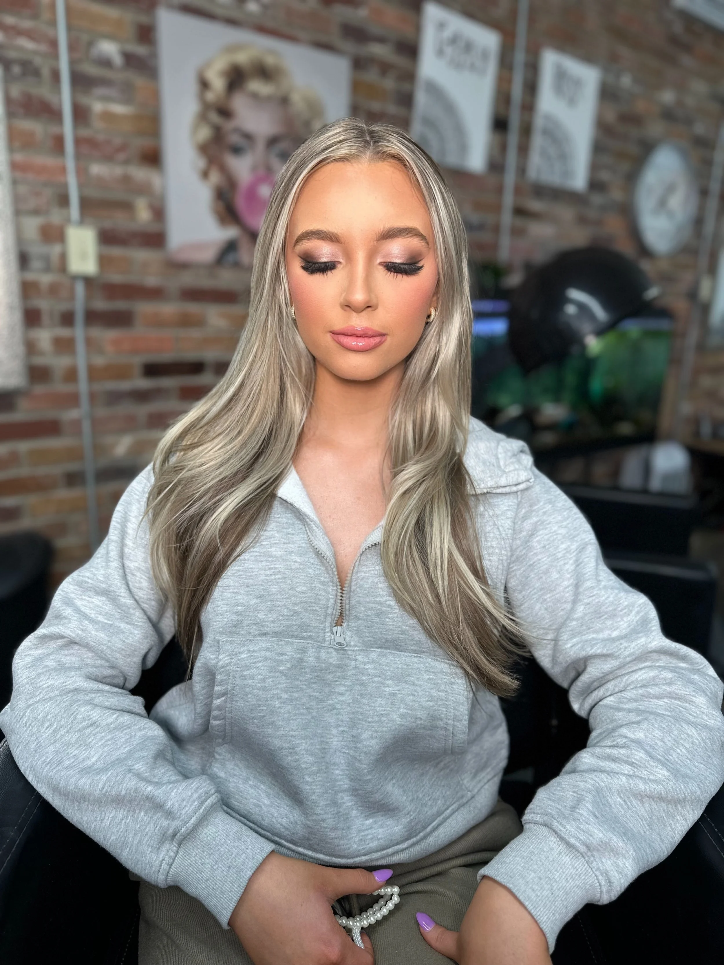 Young woman with long blonde hair, makeup, and a gray zip-up sweatshirt sitting with eyes closed in front of a brick wall with artwork.