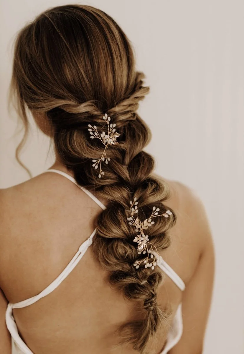 A woman with long, styled brown hair in an intricate braid adorned with decorative hairpins and floral embellishments, seen from the back.