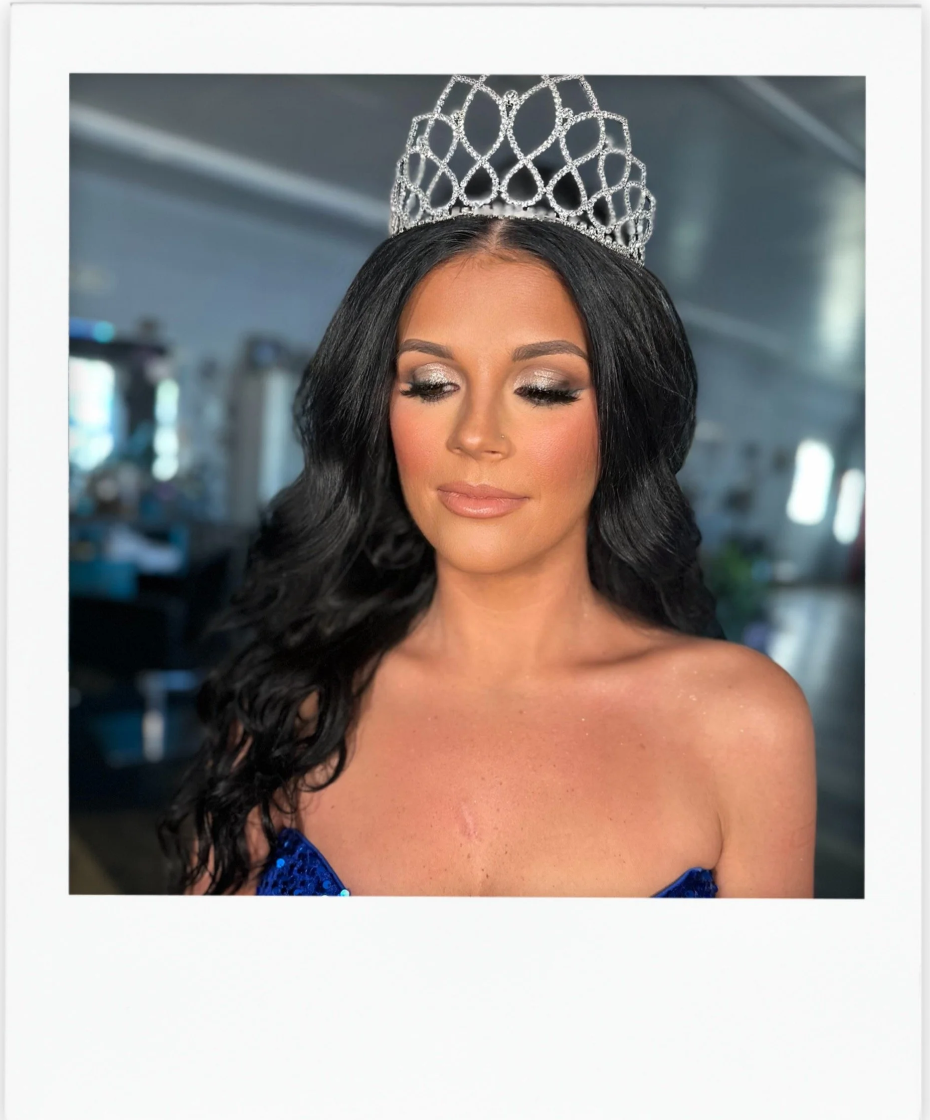 A woman with black hair wearing a silver tiara, with eyes closed and makeup, in a blue strapless dress, standing indoors with a blurred background.