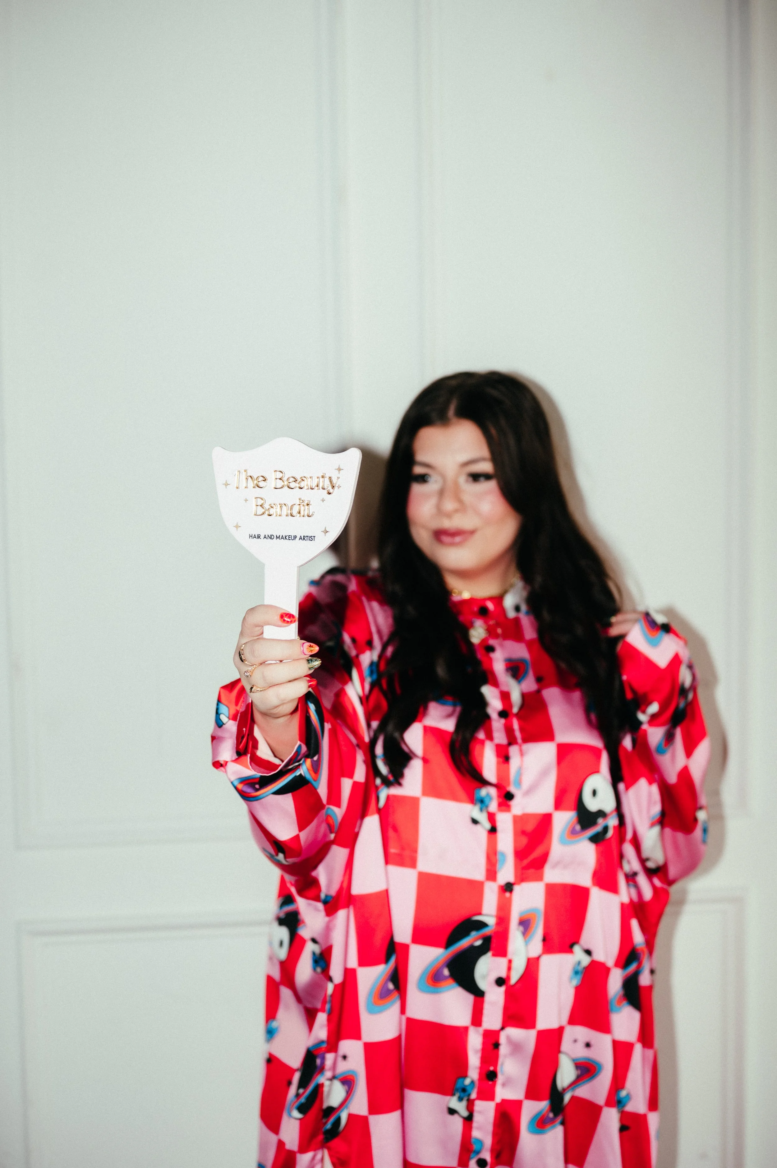 A woman with long dark hair, dressed in a colorful pink, red, and black patterned shirt, holding a fan-shaped sign that reads 'the Beauty Bandit, HAIR AND MAKEUP ARTIST.' She is standing in front of a plain white wall.