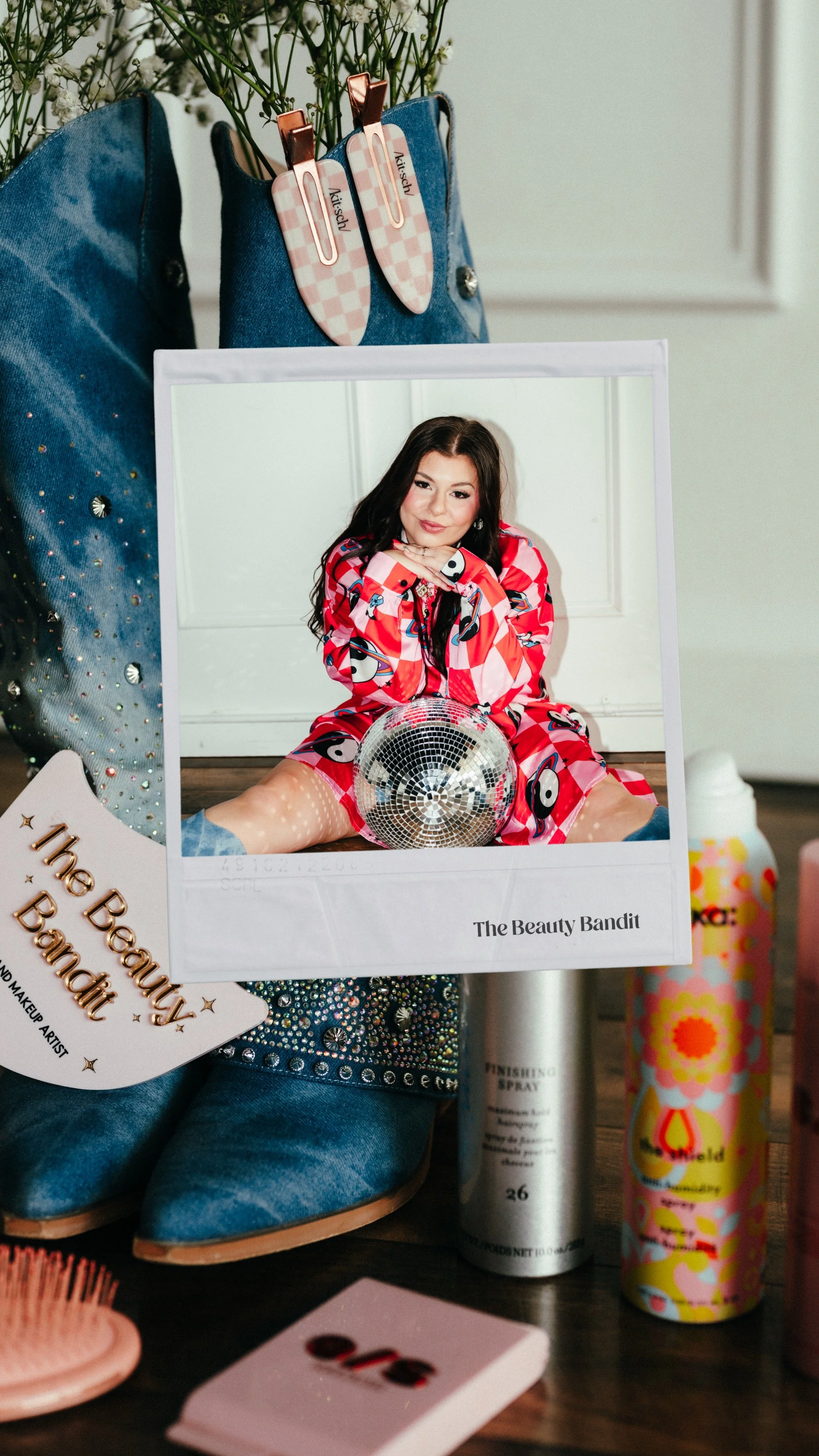 A photograph of various beauty and fashion items including a pair of blue embellished boots, a mirror with a woman in colorful clothing, a disco ball, and several bottles of spray on a wooden surface.