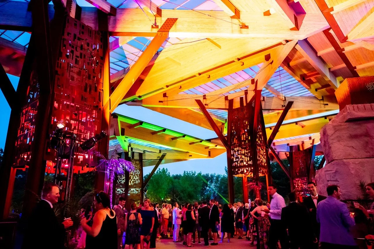 People dressed in formal attire at an indoor party under a wooden geometric ceiling with colorful LED lighting.