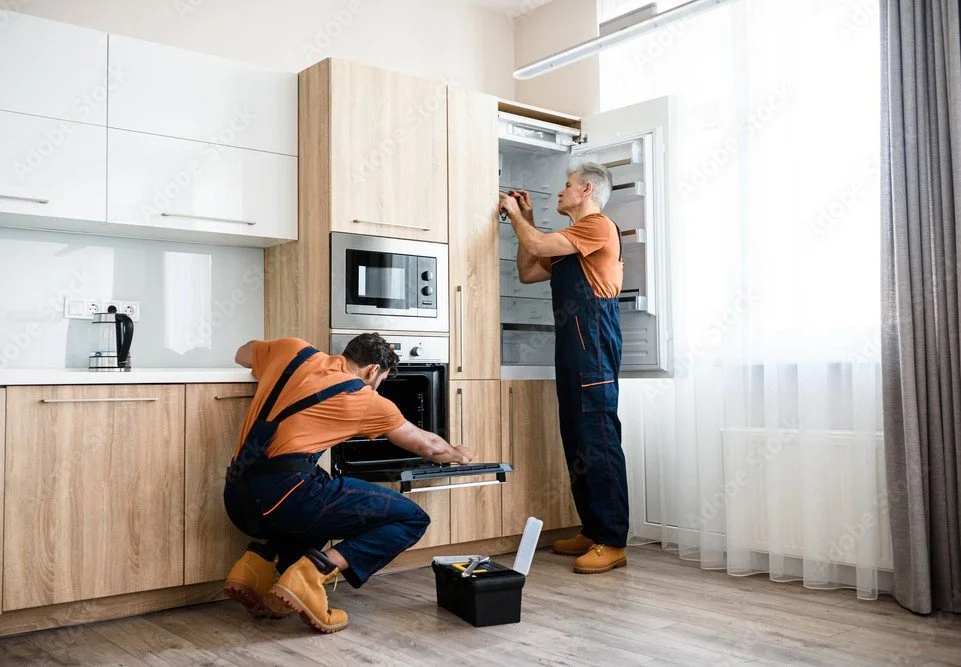 Two men in work overalls repairing an oven in a kitchen, with one opening the oven door and the other working inside the refrigerator.