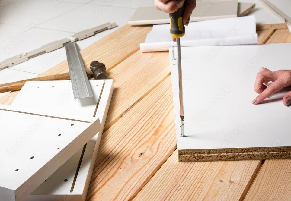Person using a screwdriver to assemble furniture with wooden and white components on a wooden surface.